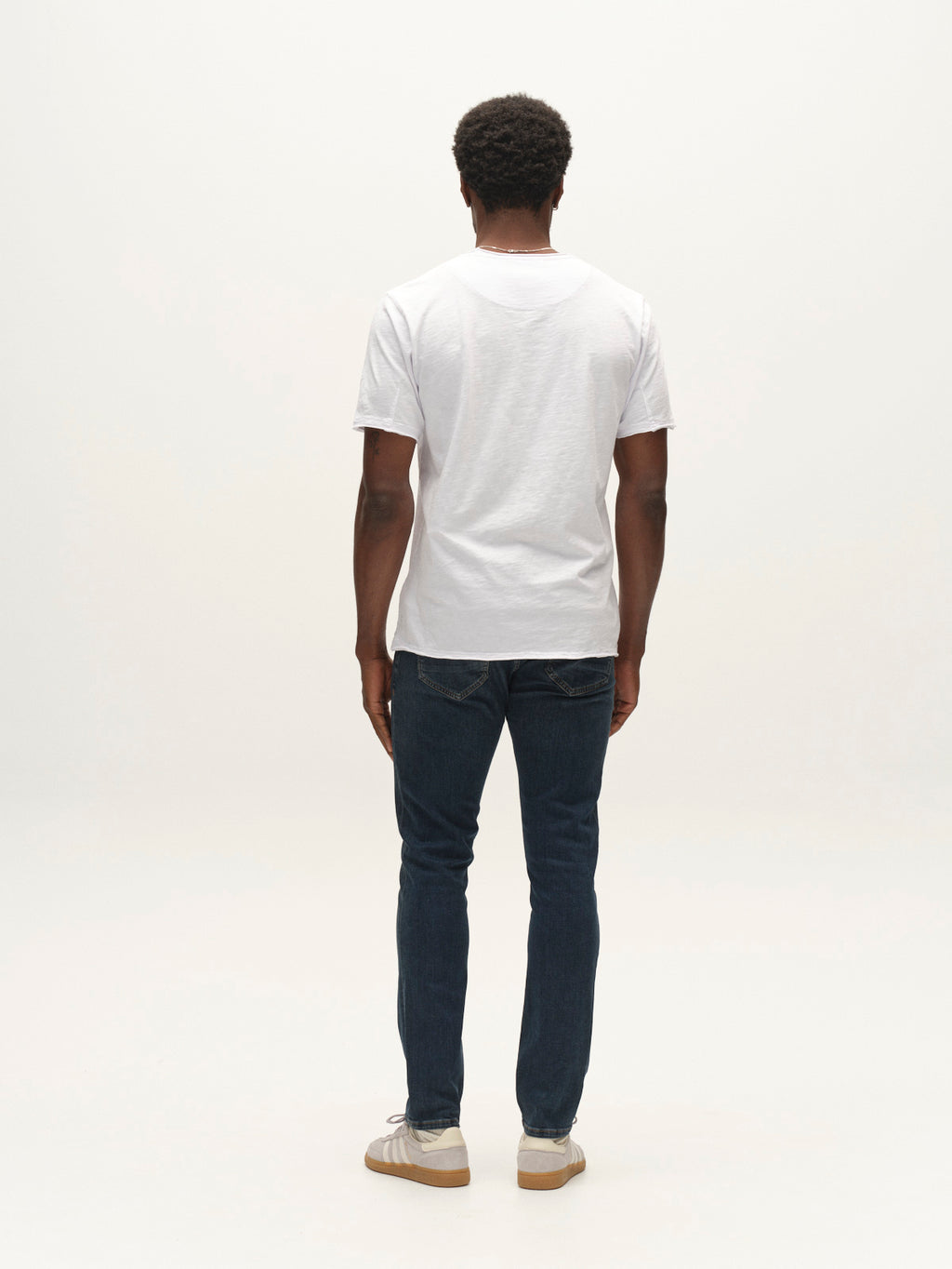 A person stands facing away from the camera, wearing a white t-shirt, Gabba GABjones2 Nham RS2113 Jeans in dark blue denim crafted from performance stretch fabric, and white sneakers against a plain, light background.