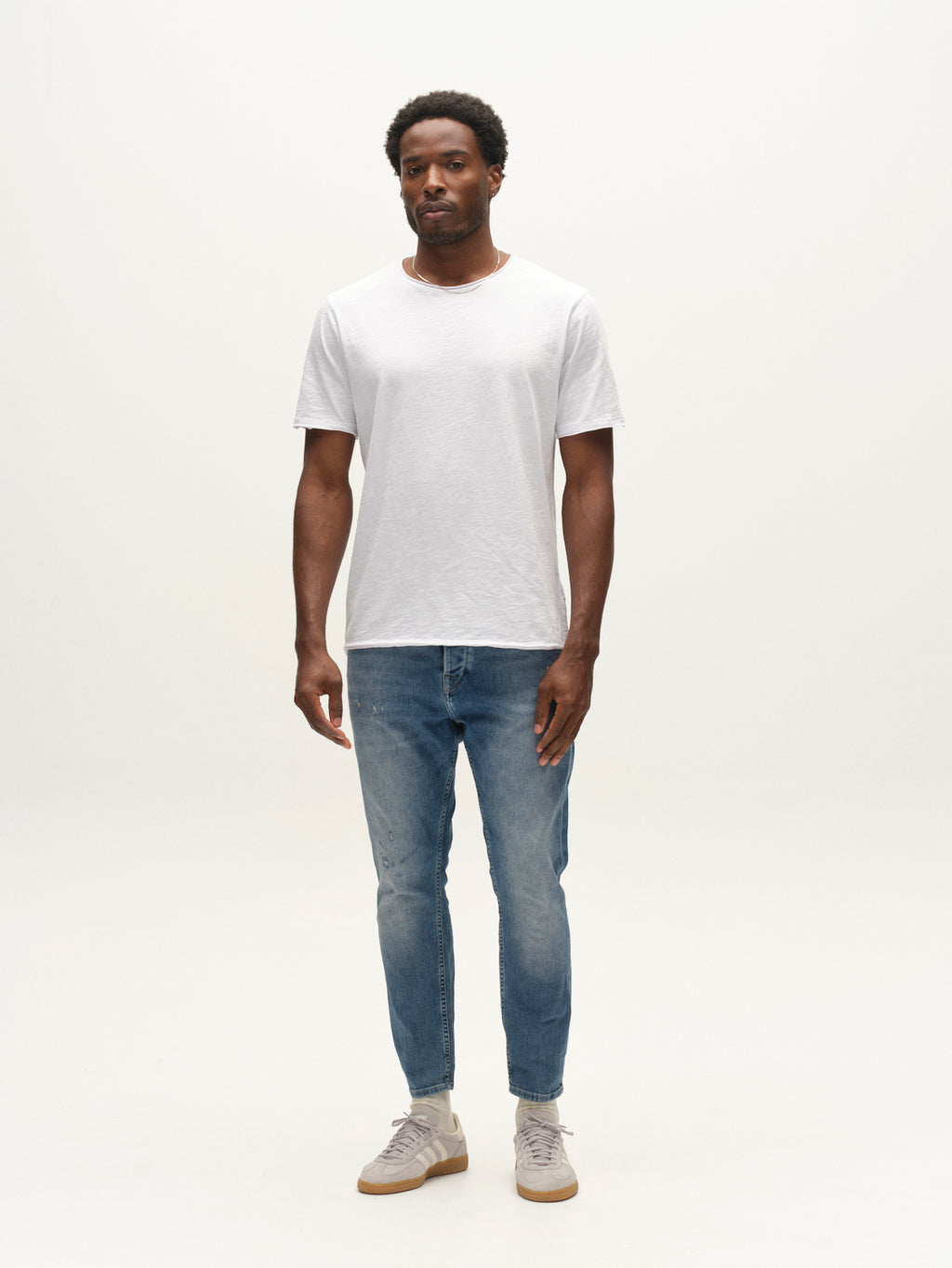 A man stands against a plain white background, wearing a white short-sleeve t-shirt, light gray sneakers, and Gabba GABalex Cropped Nham RS2112 Jeans in mid blue denim. The jeans are crafted with ISKO stretch denim for performance comfort. He faces forward with a neutral expression and relaxed posture.