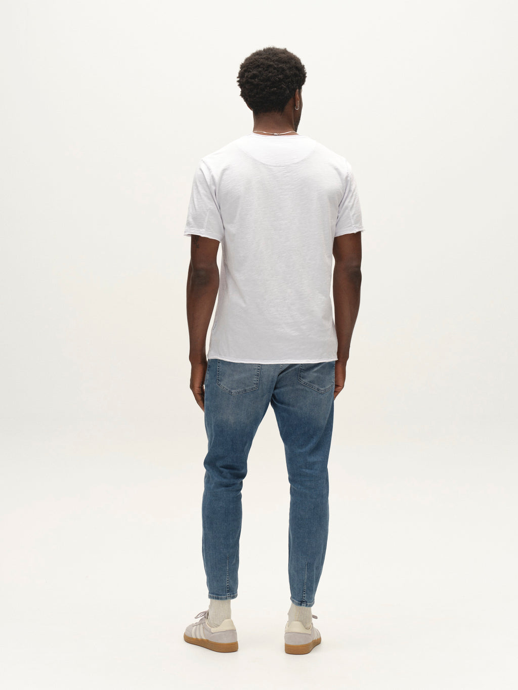 A person stands facing away from the camera, wearing a plain white t-shirt, Gabba GABalex Cropped Nham RS2112 Jeans in mid blue denim crafted from ISKO stretch denim for enhanced performance comfort, and beige sneakers, set against a simple white background.