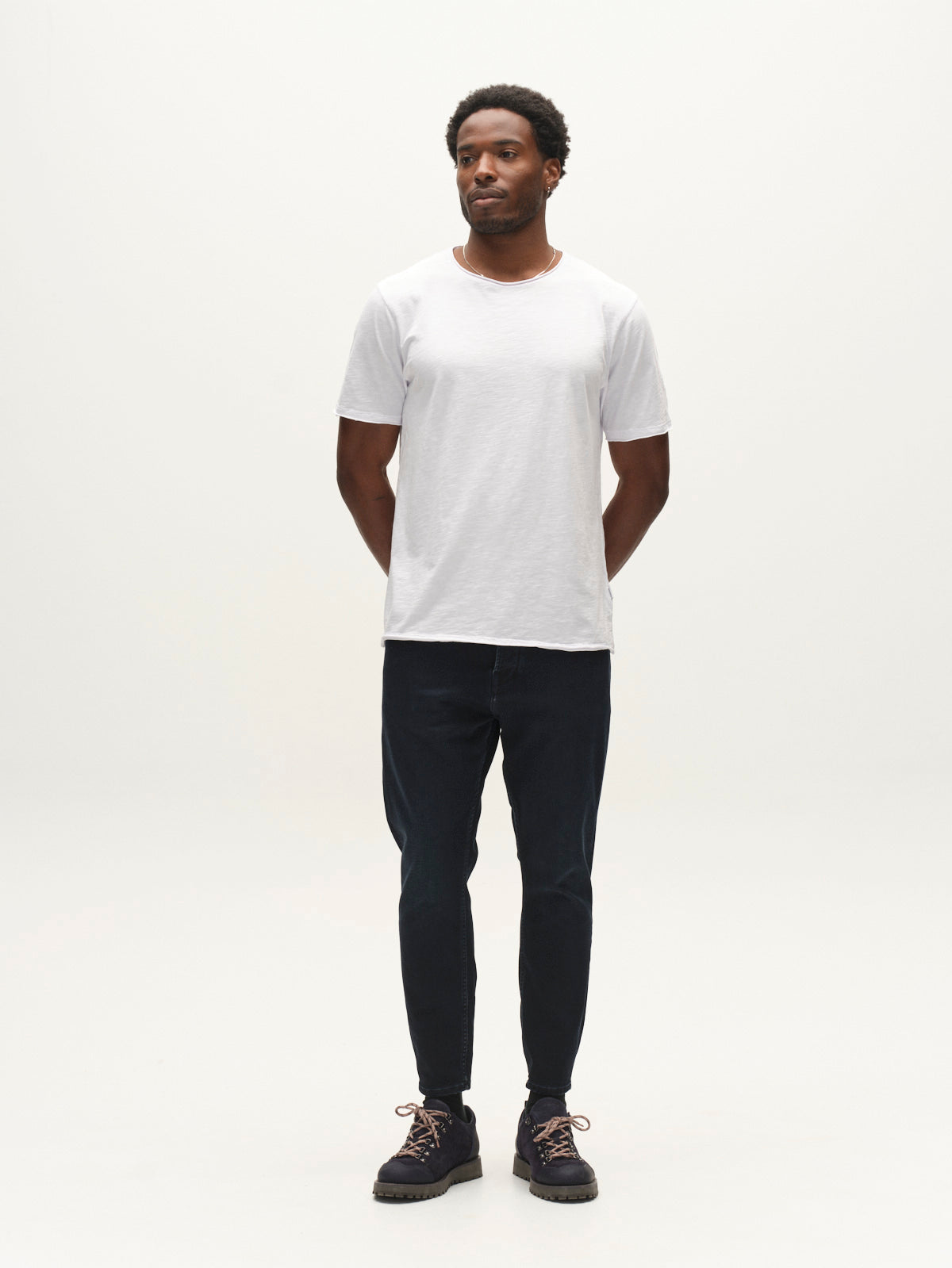 A man stands in a minimal studio, wearing a white t-shirt, Gabba GABalex Cropped Torino RS2106 Jeans in dark blue denim with a relaxed tapered fit, and black shoes. His hands are behind his back as he looks slightly to the side. The background is plain white.