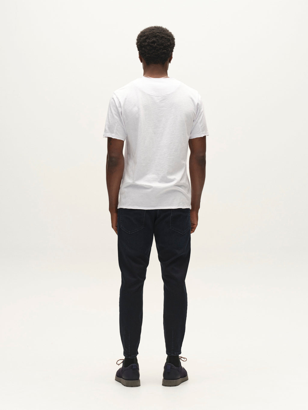 A man stands facing away from the camera, wearing a plain white T-shirt, black shoes, and Gabba GABalex Cropped Torino RS2106 Jeans in dark blue denim with a relaxed tapered fit, against a plain white background.