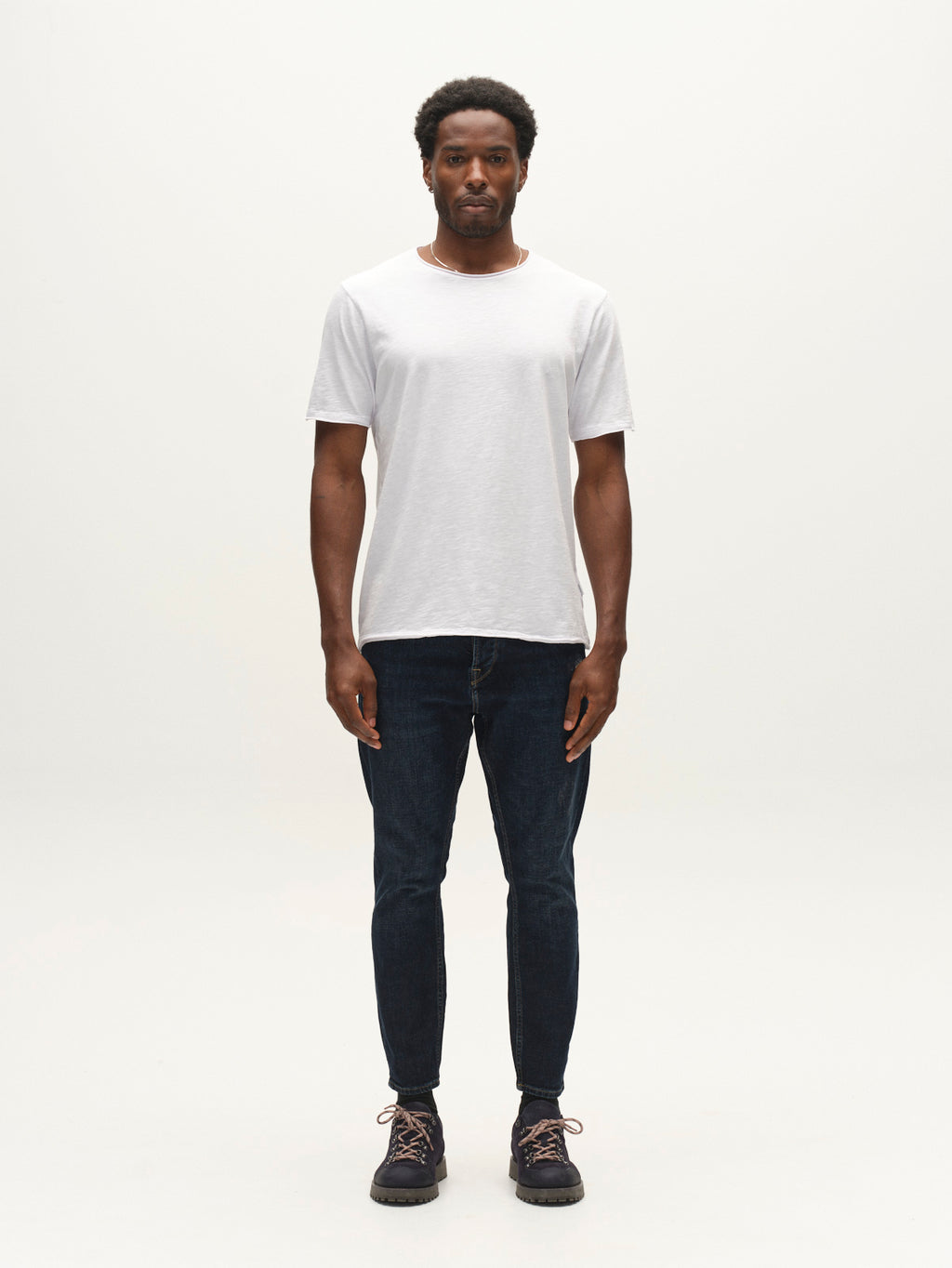 A man stands facing forward in a plain white t-shirt, Gabba GABalex Cropped Cros RS2108 Jeans RCS in dark blue denim with a relaxed tapered fit, and black sneakers against a simple light background.