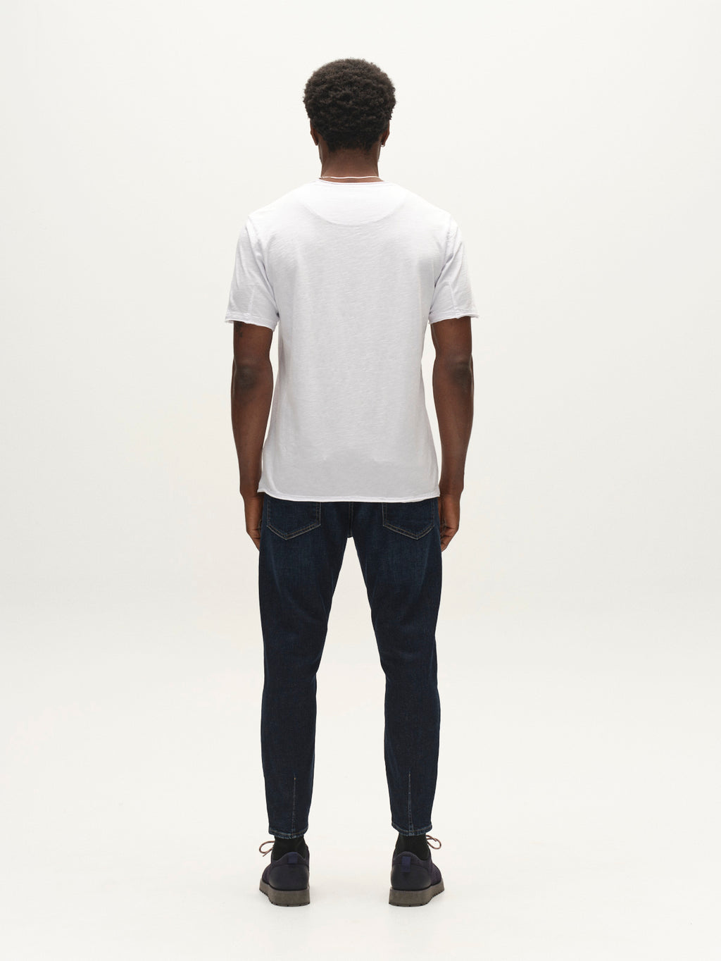 A person stands facing away, wearing a plain white t-shirt, dark shoes, and Gabba GABalex Cropped Cros RS2108 Jeans RCS in Dark Blue Denim with a relaxed tapered fit, against a plain light background.