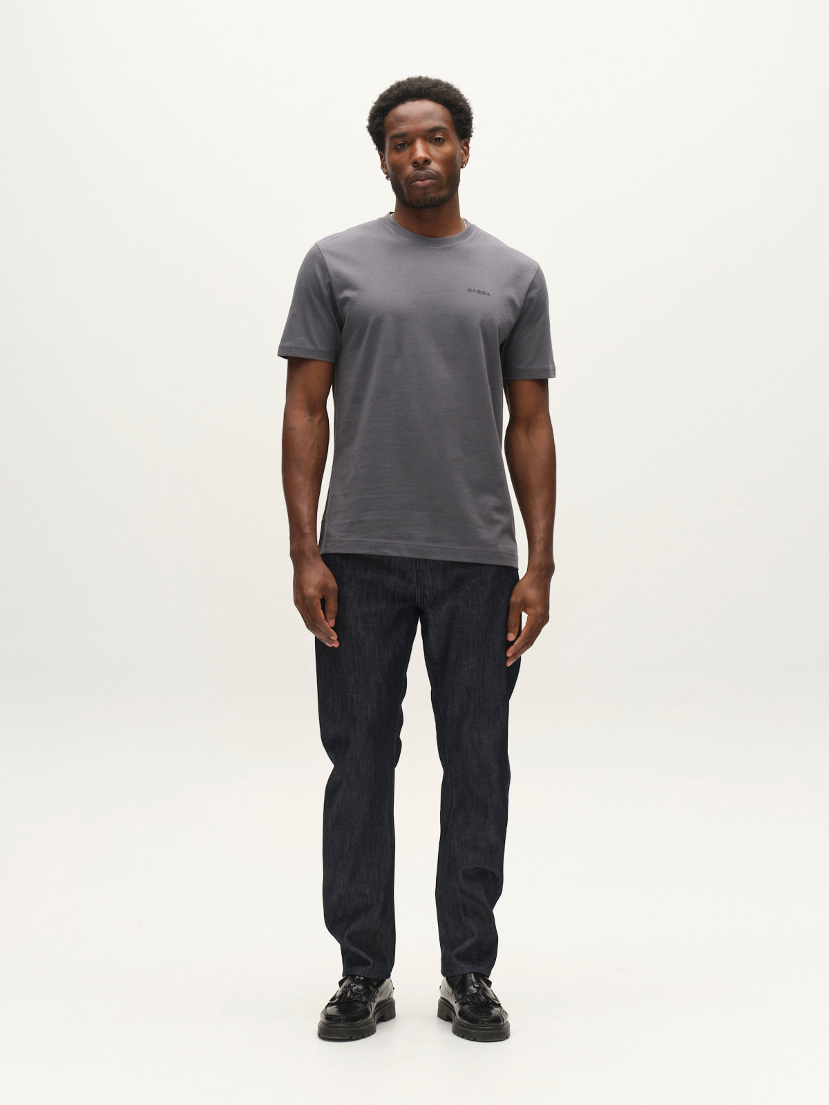 A man stands facing forward against a plain white background, wearing a gray short-sleeve t-shirt, black shoes, and Gabba GABcarl Anark Stone Raw Jeans in dark blue denim with a loose tapered fit.