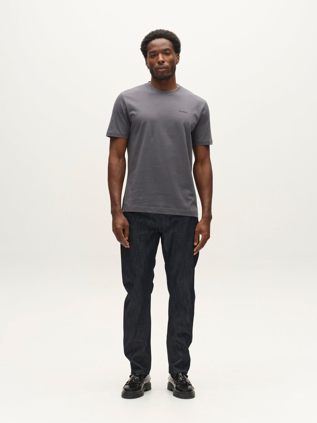 A man stands facing forward against a plain white background, wearing a gray short-sleeve t-shirt, black shoes, and Gabba GABcarl Anark Stone Raw Jeans in dark blue denim with a loose tapered fit.