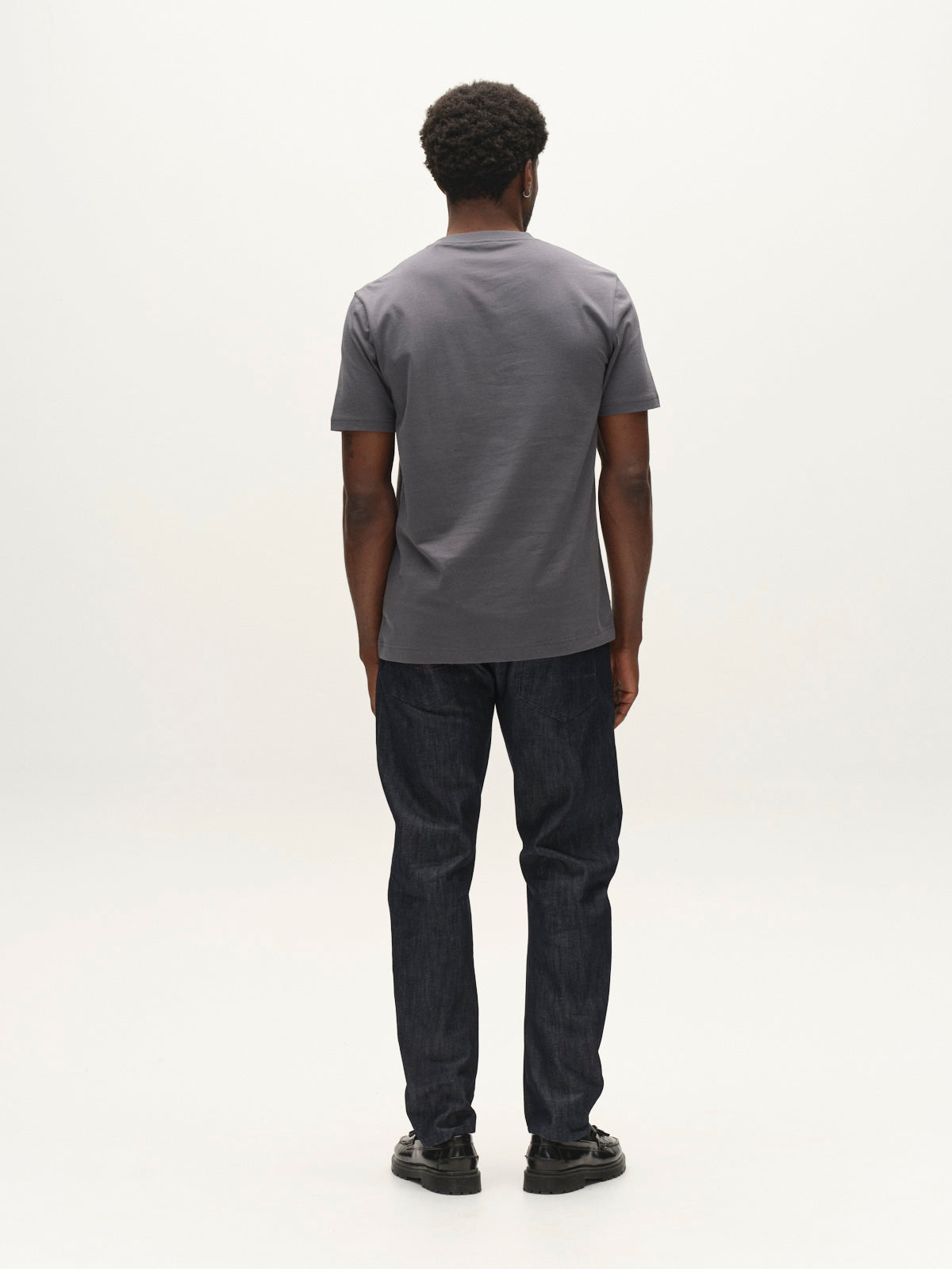 A man stands facing away from the camera, wearing a gray t-shirt, Gabba GABcarl Anark Stone Raw Jeans in dark blue denim, featuring a loose tapered fit, and black shoes, against a plain white background.
