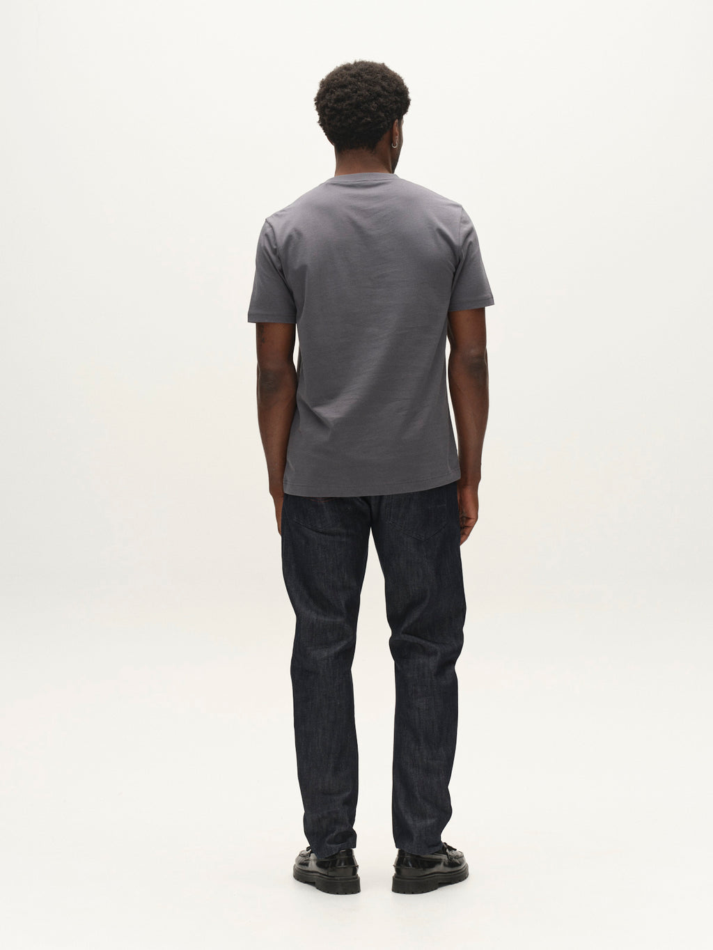 A man stands facing away from the camera, wearing a gray t-shirt, Gabba GABcarl Anark Stone Raw Jeans in dark blue denim, featuring a loose tapered fit, and black shoes, against a plain white background.