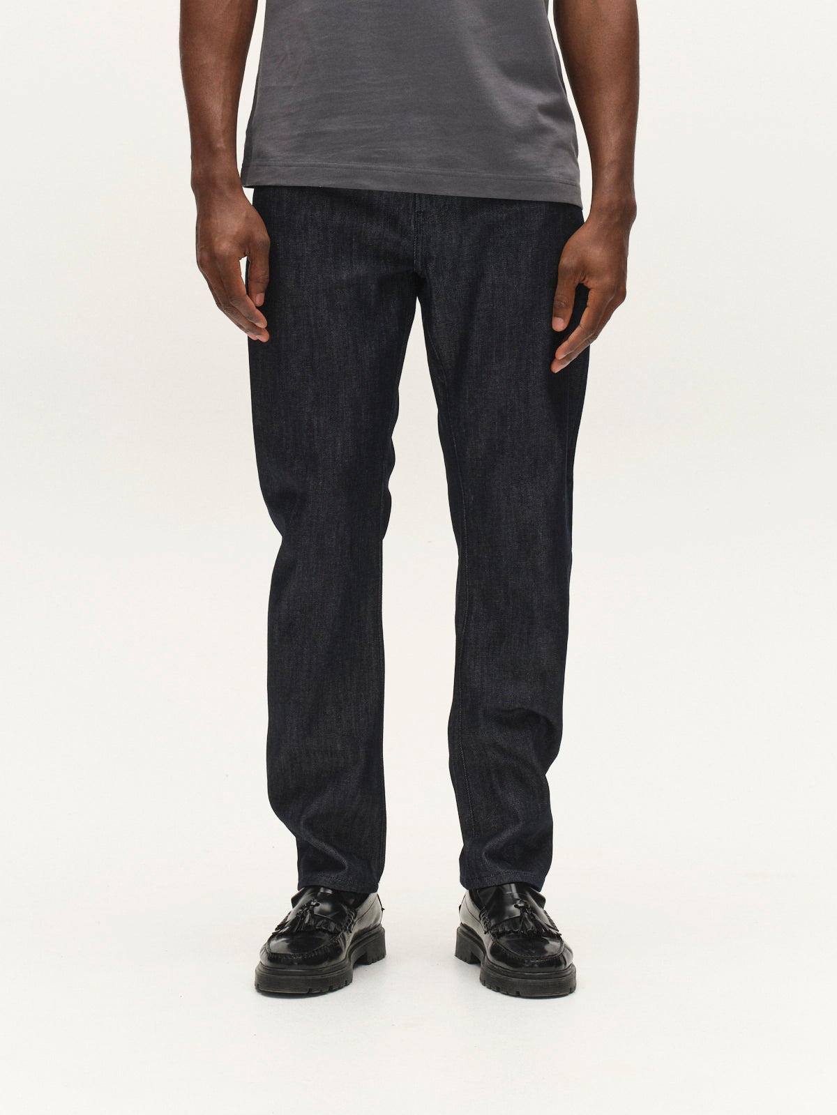 A person stands against a plain white background, their upper body cropped out from the shoulders down. They are wearing a gray t-shirt, black shoes, and Gabba GABcarl Anark Stone Raw Jeans in dark blue denim—loose tapered fit jeans crafted from premium ISKO denim.