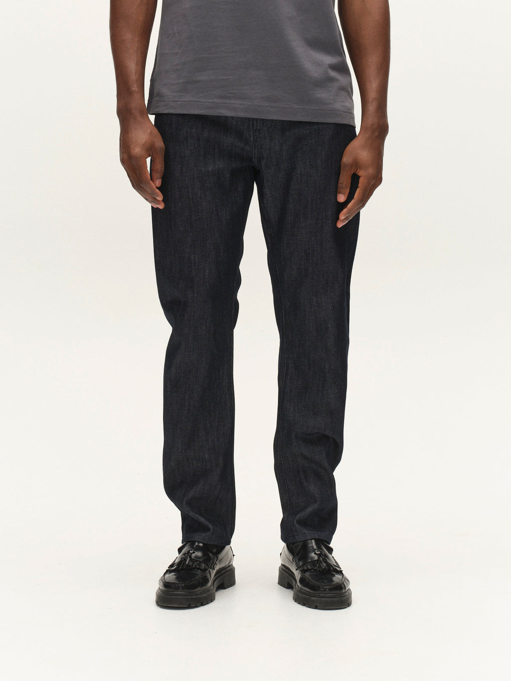 A person stands against a plain white background, their upper body cropped out from the shoulders down. They are wearing a gray t-shirt, black shoes, and Gabba GABcarl Anark Stone Raw Jeans in dark blue denim—loose tapered fit jeans crafted from premium ISKO denim.