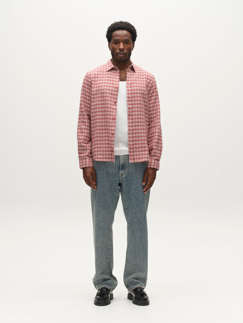 A man stands facing forward against a plain white background, wearing a pink plaid shirt layered over a white t-shirt, paired with Gabba GABnoah Tona RS2100 Jeans in mid blue denim—crafted from rigid indigo fabric in a loose straight fit—and black shoes.