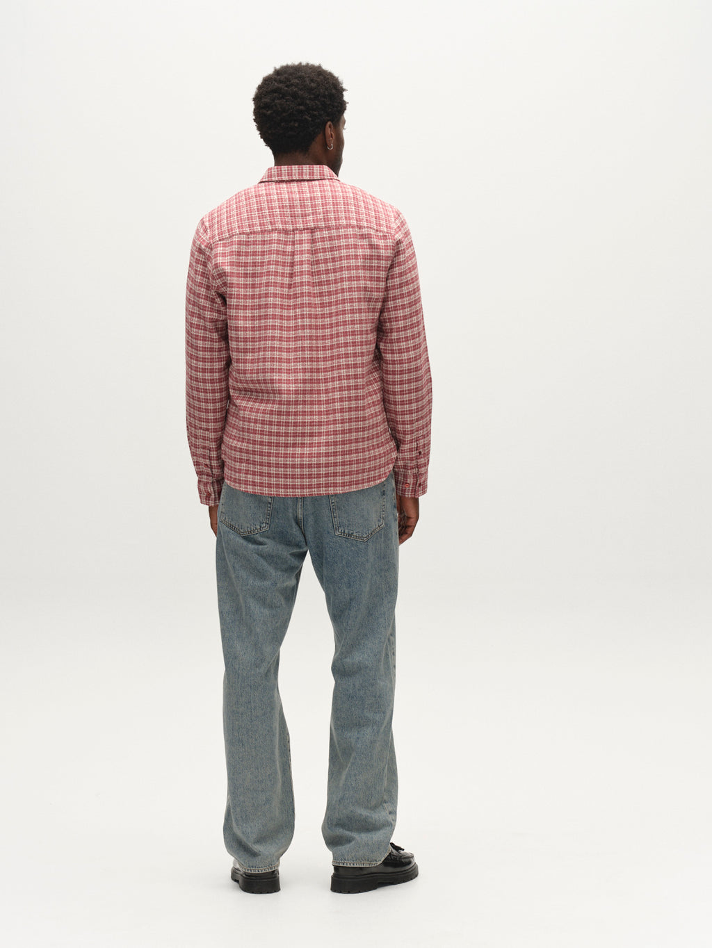 A person stands facing away, wearing a red and white plaid long-sleeve shirt, Gabba GABnoah Tona RS2100 Jeans in mid blue denim, and black shoes, against a plain white background.