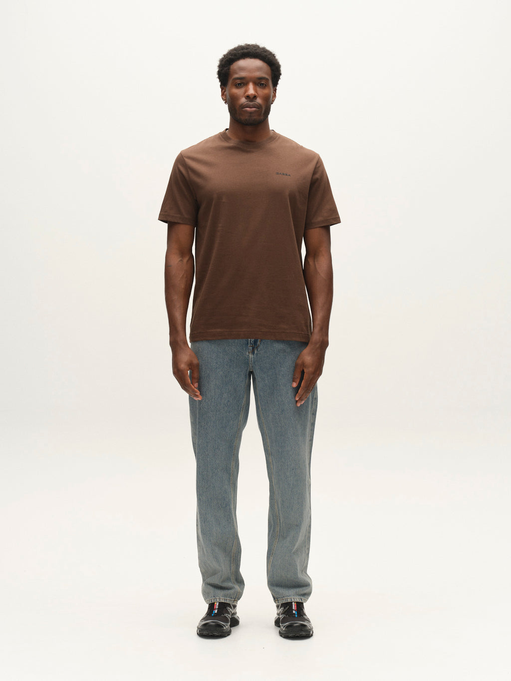 A man stands facing forward against a plain light background, wearing a brown short-sleeve T-shirt, Gabba GABmax Tona RS2100 Jeans in mid blue denim, and black shoes.