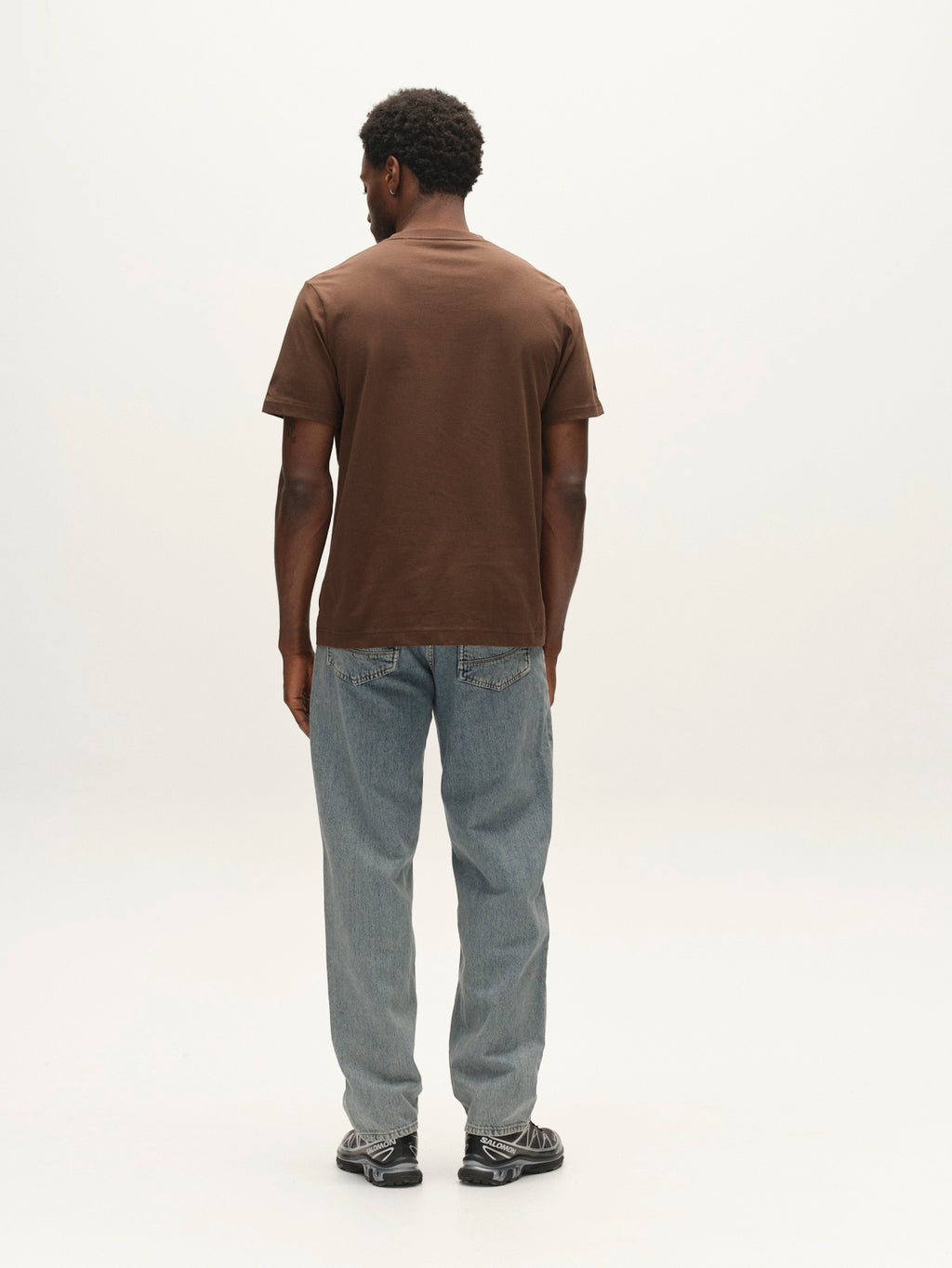 A person stands facing away, wearing a brown short-sleeve shirt, Gabba GABmax Tona RS2100 Jeans in mid blue denim crafted from organic cotton, and black sneakers, against a plain white background.