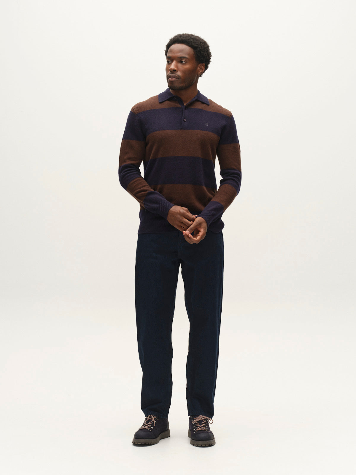 A man stands against a plain light background wearing a dark brown and navy striped sweater, Gabba GABsenna Denia Rinse Jeans in dark blue denim, and brown shoes, looking slightly to the side with his hands loosely clasped.