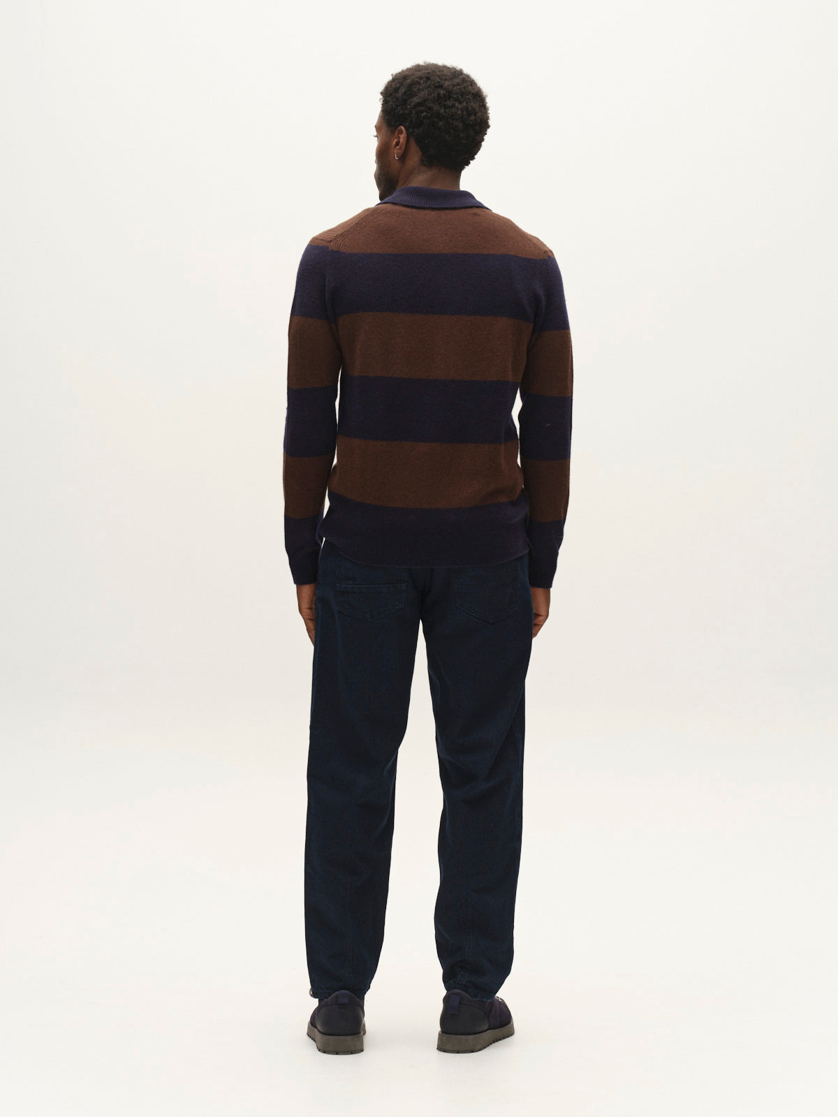 A man stands facing away, wearing a brown and navy striped sweater, Gabba GABsenna Denia Rinse Jeans in dark blue denim, and dark shoes against a plain white background.