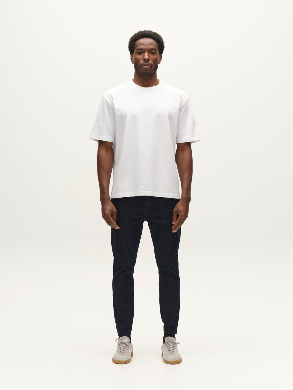A man stands facing forward in a plain studio, wearing a white t-shirt, light-colored sneakers, and the Gabba GABalex Cropped Bekham DS0001 Jeans in dark blue denim. The background is simple and uncluttered.