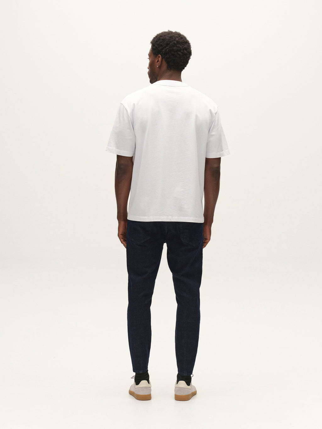 A man stands facing away from the camera, wearing a plain white T-shirt, beige sneakers, and Gabba GABalex Cropped Bekham DS0001 Jeans in dark blue denim with a relaxed tapered fit crafted from comfort stretch fabric, against a plain white background.