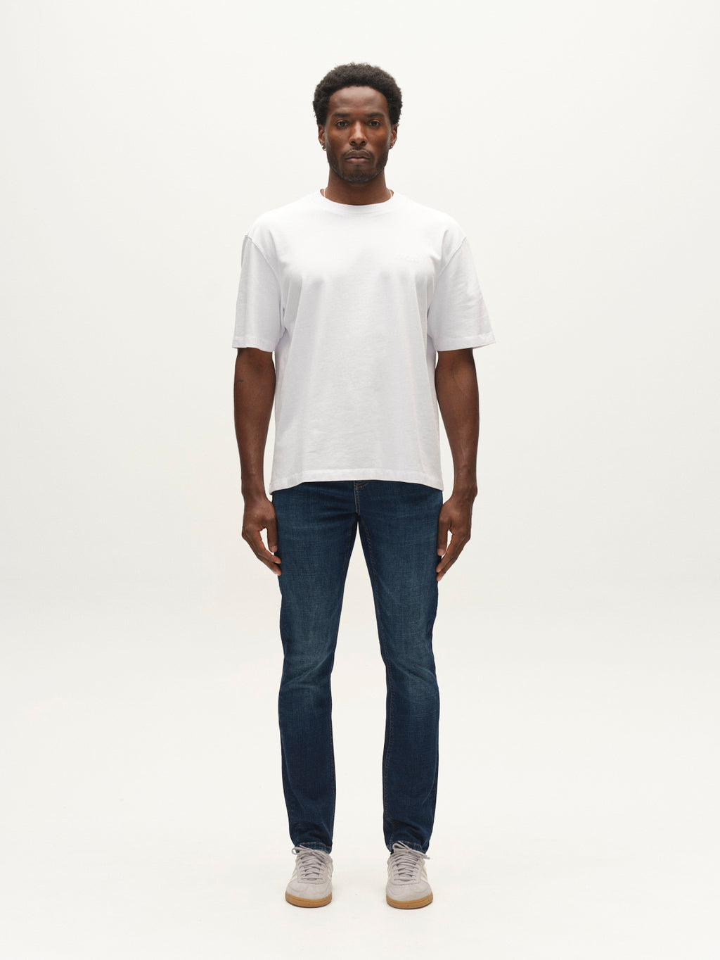 A man stands facing forward against a plain white background, wearing a white t-shirt, gray sneakers, and Gabba GABjones Bekham DS0002 Jeans in dark blue denim.