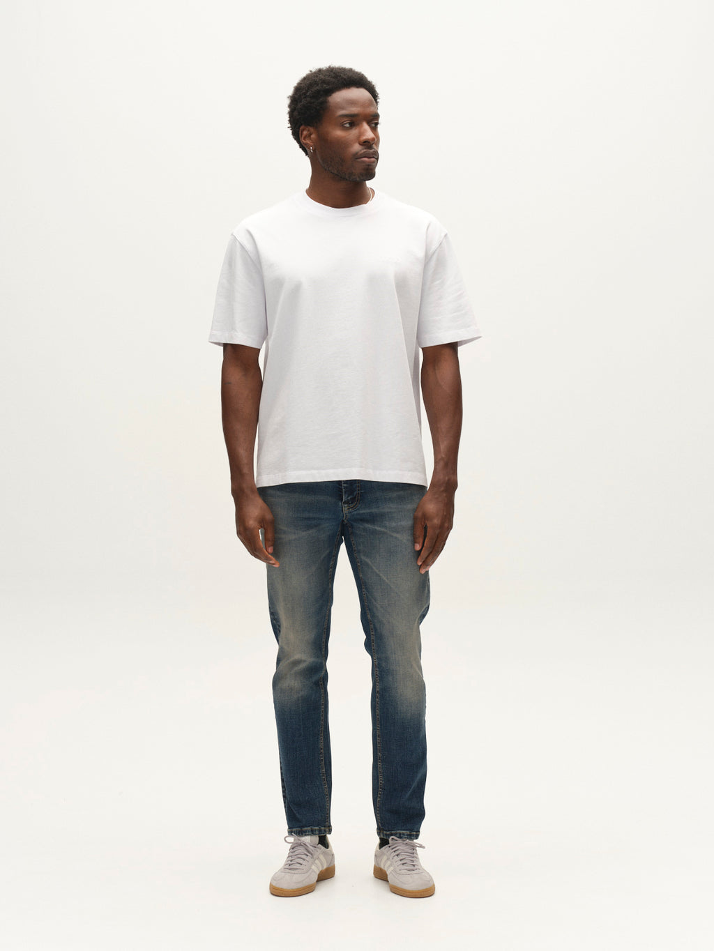 A man stands against a plain white background wearing a regular fit white t-shirt, Gabba GABmarc Bekham DS0003 Jeans in mid-blue denim, and light gray sneakers, looking slightly to his left with a neutral expression.