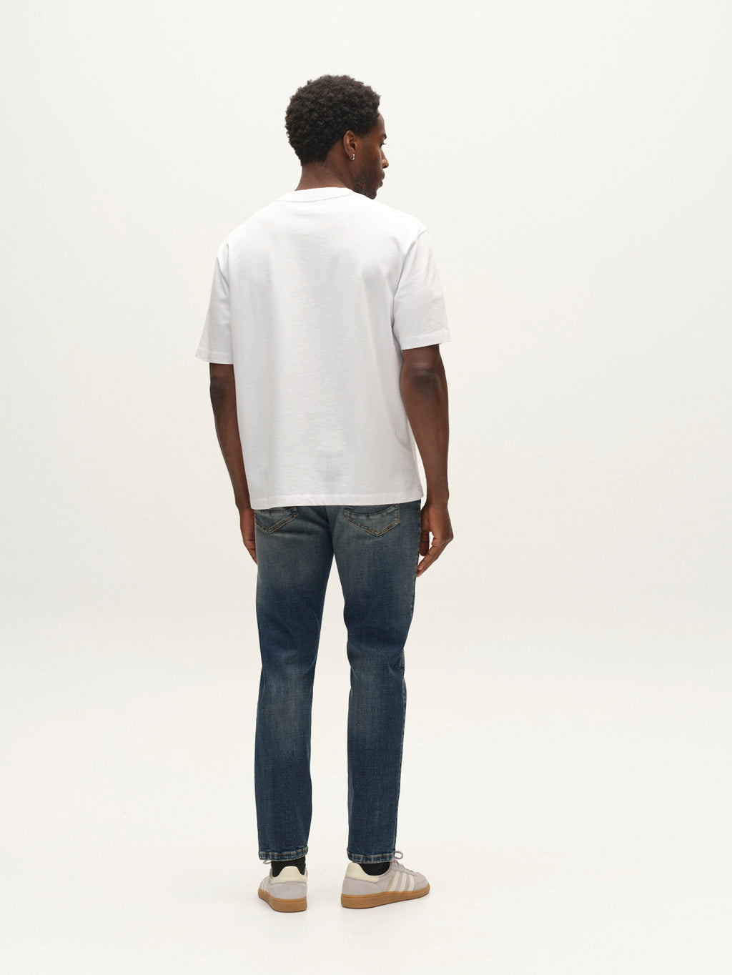 A man stands facing away, wearing a plain white t-shirt and beige sneakers, paired with Gabba GABmarc Bekham DS0003 Jeans in mid-blue denim crafted from comfort stretch fabric, against a plain light background.