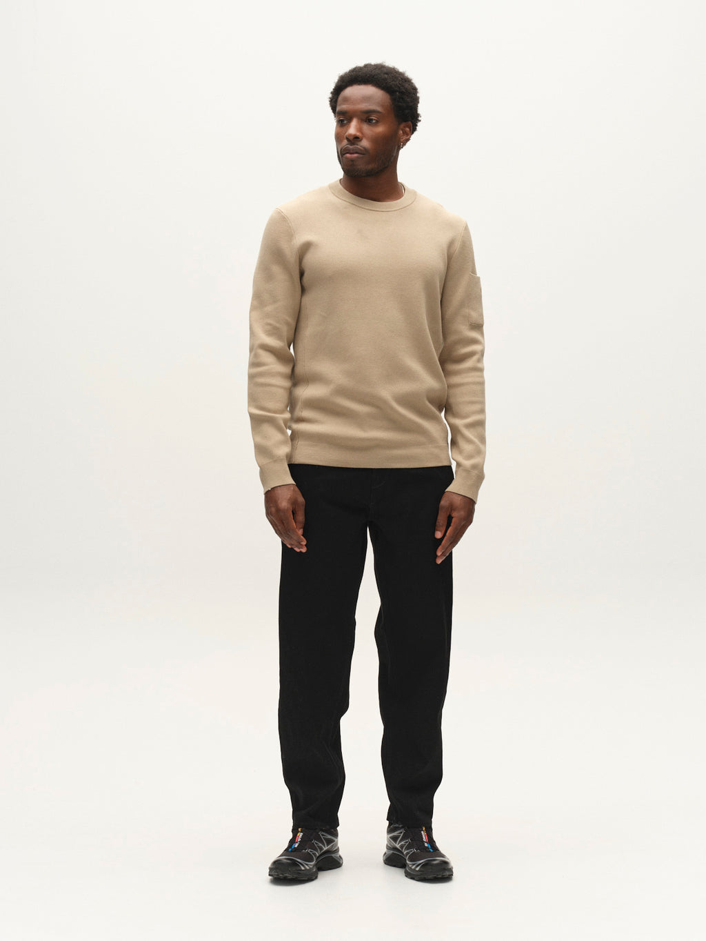 A man stands against a plain white background, wearing a beige sweater, Gabba GABsenna Black DS0006 Jeans in black denim, and black shoes. He faces slightly to the side with his hands relaxed by his sides.