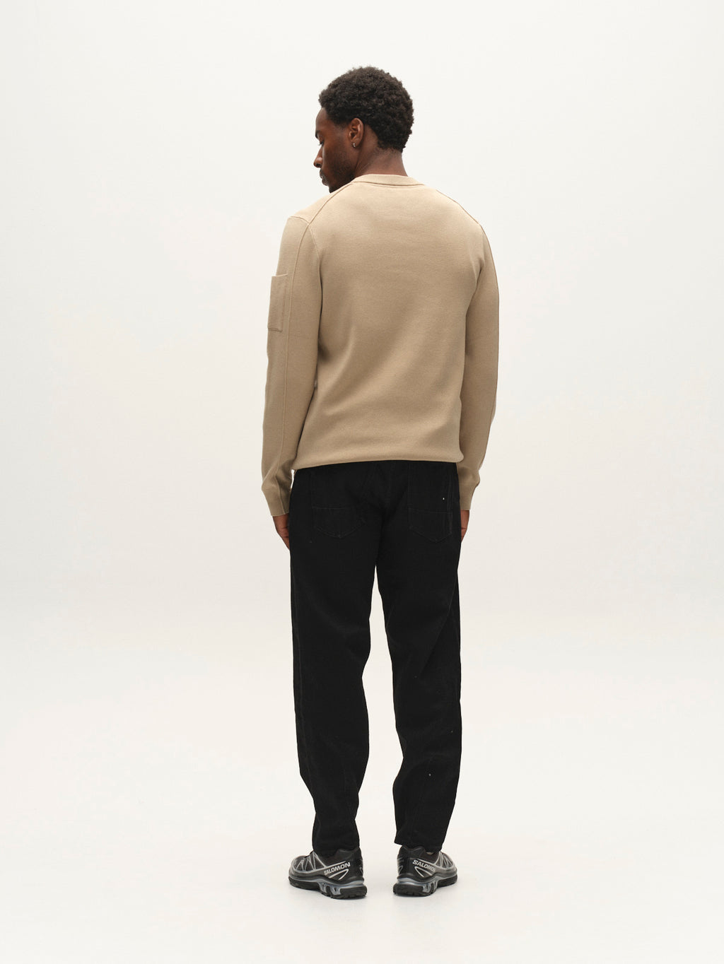 A man stands facing away, wearing a beige long-sleeve shirt and the Gabba GABsenna Black DS0006 Jeans in black denim, which feature a loose tapered fit. He’s paired them with black sneakers with white soles, set against a plain light-colored background.