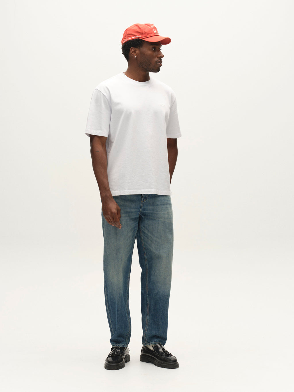 A man stands against a plain background, wearing a red cap, a white t-shirt, Gabba GABsenna Wolv DS0004 Jeans in dark blue denim, and black shoes. He is looking to his left with his arms relaxed by his sides.