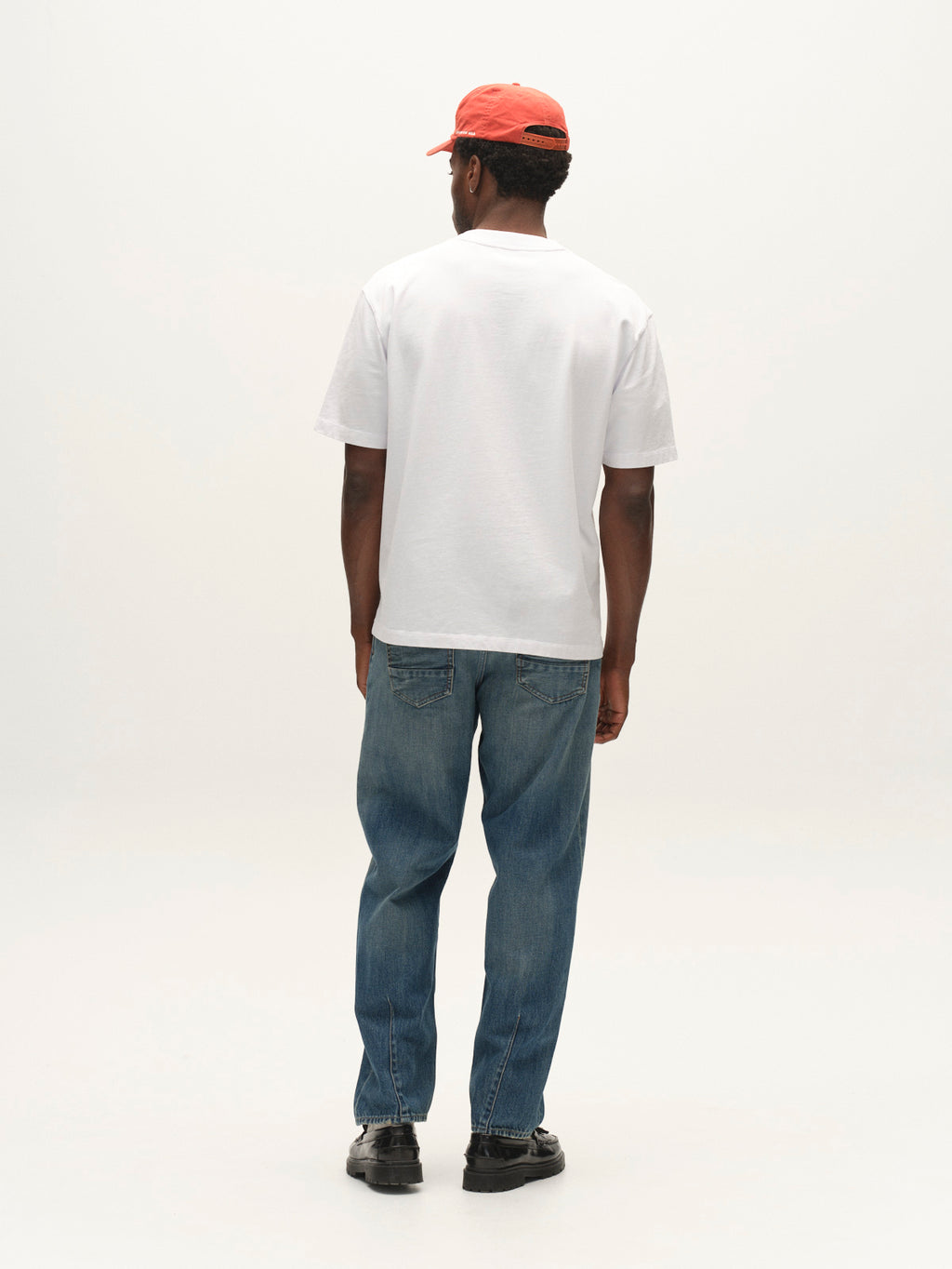 A man stands facing away, wearing a white t-shirt, Gabba GABsenna Wolv DS0004 Jeans in Dark Blue Denim, black shoes, and a red cap. The background is plain and light.