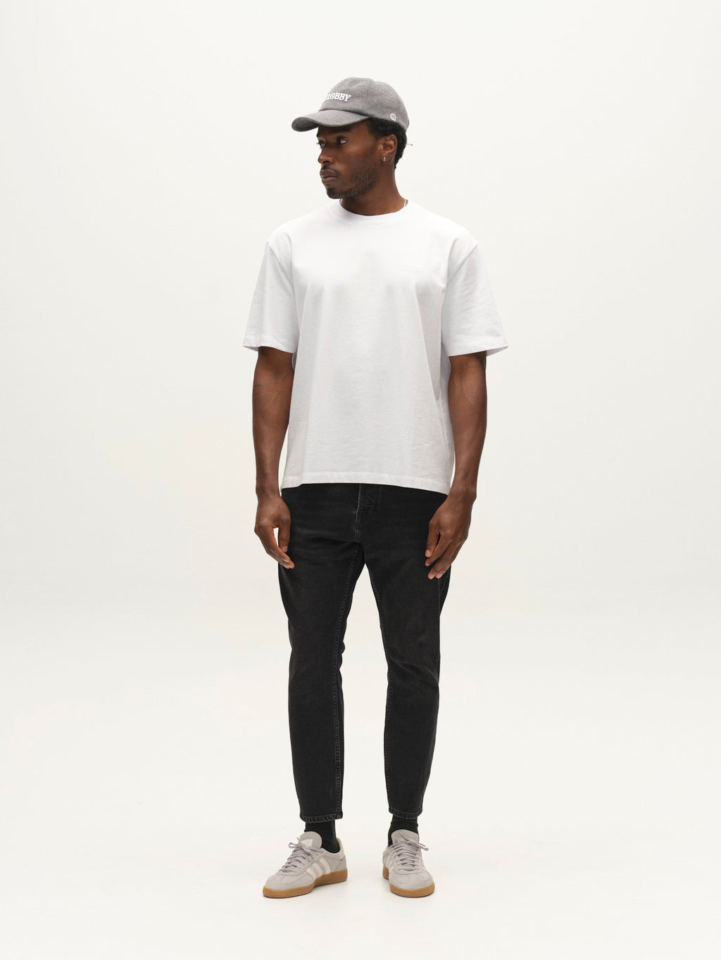 A man wearing a gray cap, white t-shirt, and gray sneakers stands facing slightly to the right against a plain white background. He completes his effortlessly stylish look with Gabba’s GABalex Cropped Blerb WW0031 Jeans in black denim, featuring a relaxed tapered fit crafted from high-density fabric.