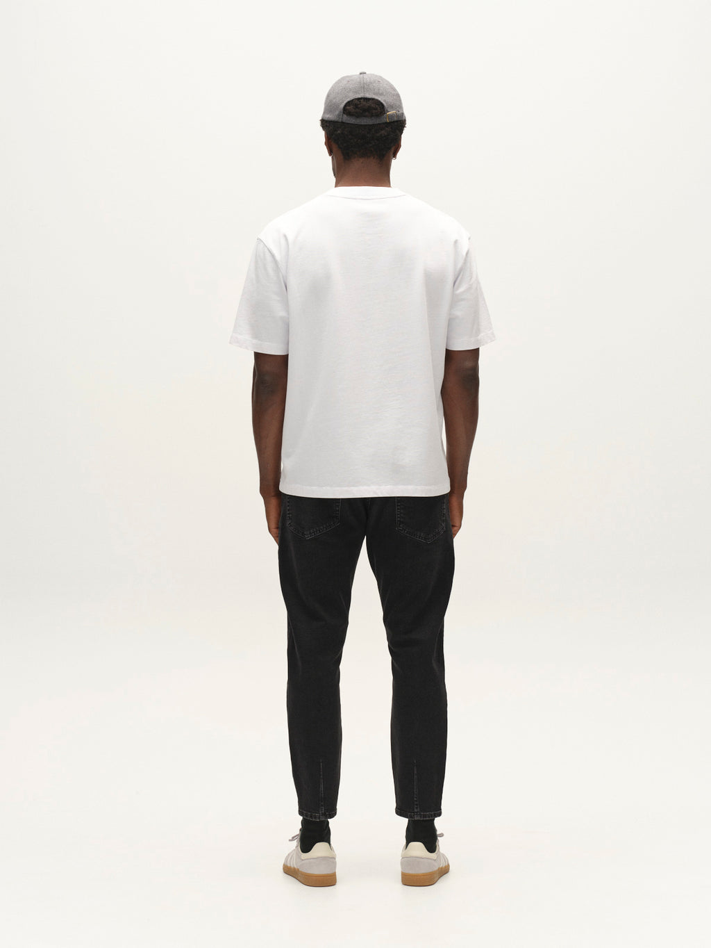 A person stands facing away from the camera, wearing a gray cap, a white t-shirt, and Gabba GABalex Cropped Blerb WW0031 Jeans in black denim—featuring a relaxed tapered fit crafted from high-density fabric—paired with white sneakers against a plain white background.