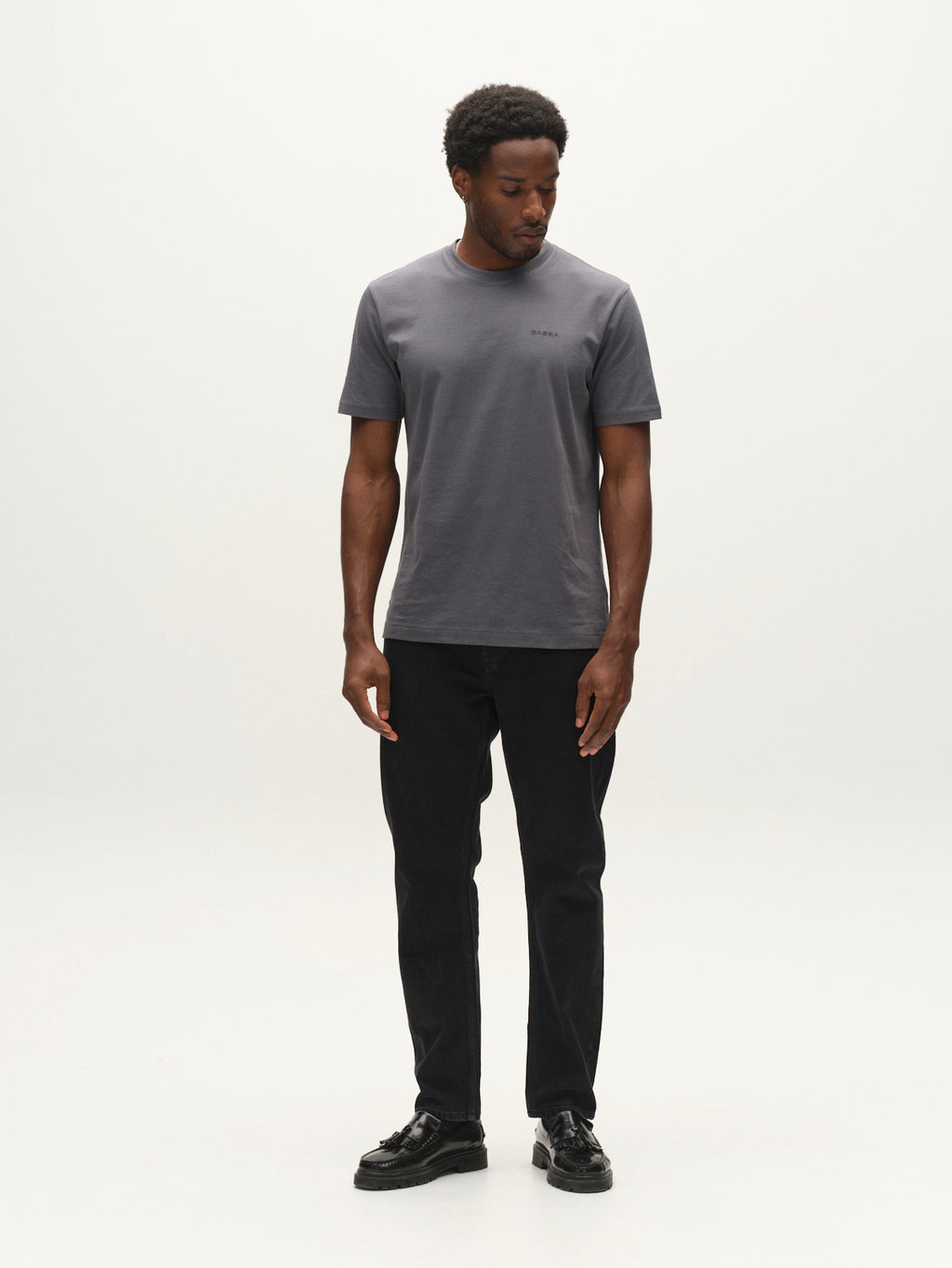 A man stands in a minimal studio wearing a gray t-shirt, Gabba GABcarl Blaccom WW0035 Jeans in black denim with a loose tapered fit, and black shoes. He looks down and to his left with his arms relaxed by his sides.
