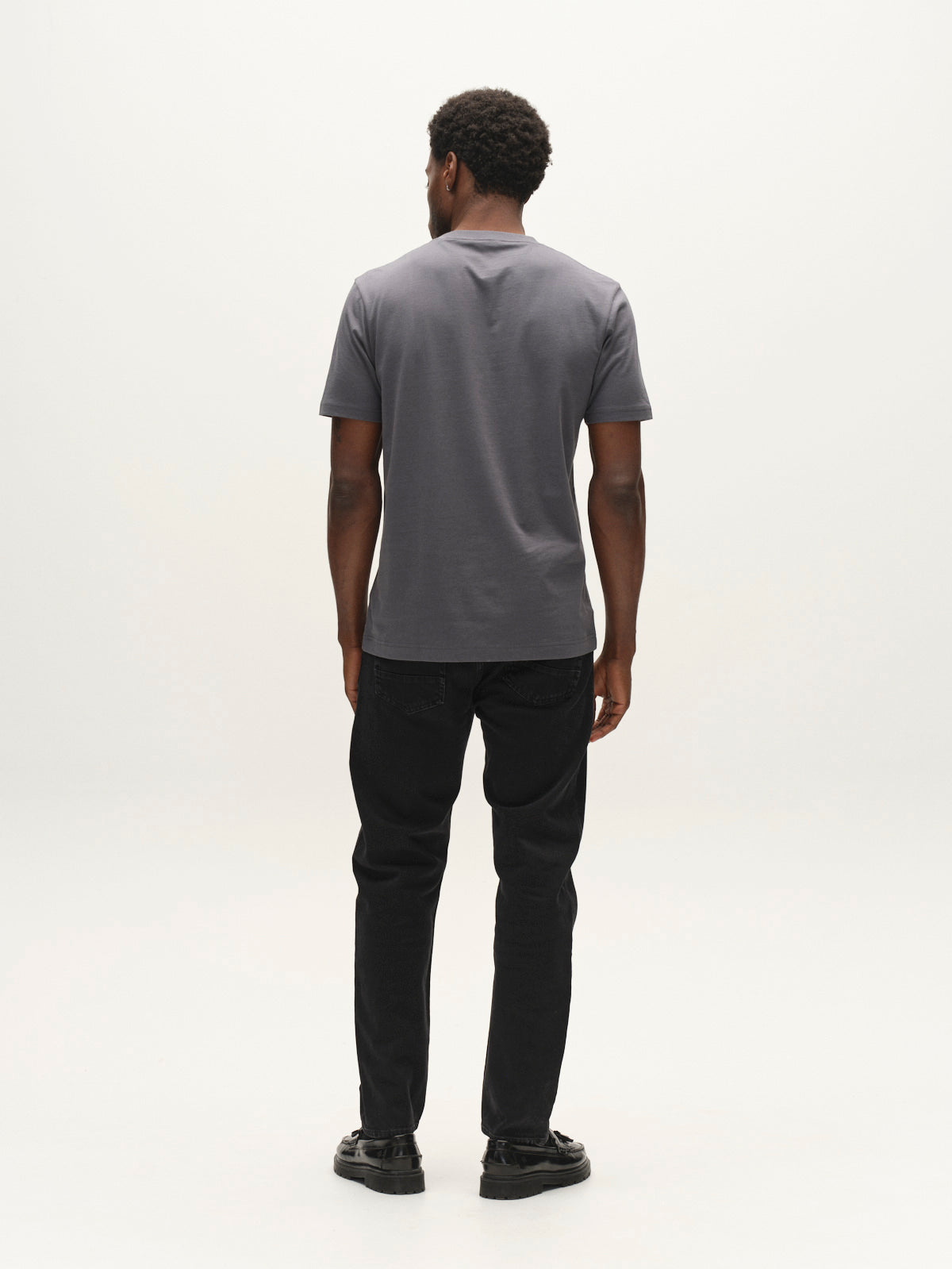 A man stands facing away from the camera, wearing a short-sleeved gray t-shirt, black shoes, and Gabba GABcarl Blaccom WW0035 Jeans in Black Denim with a loose tapered fit, against a plain white background.