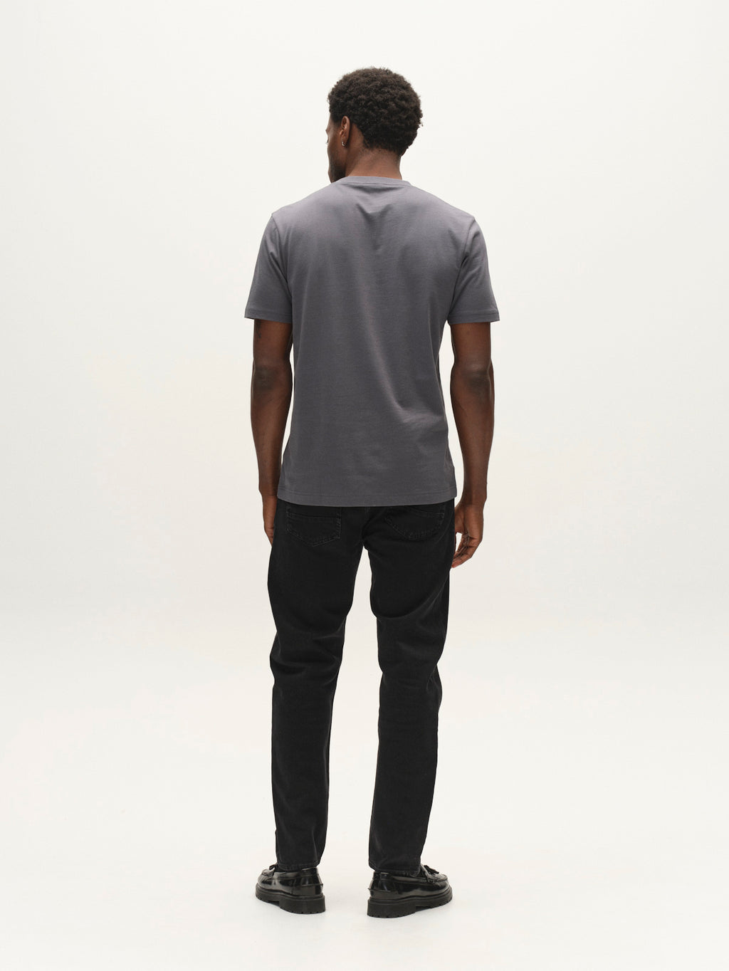 A man stands facing away from the camera, wearing a short-sleeved gray t-shirt, black shoes, and Gabba GABcarl Blaccom WW0035 Jeans in Black Denim with a loose tapered fit, against a plain white background.