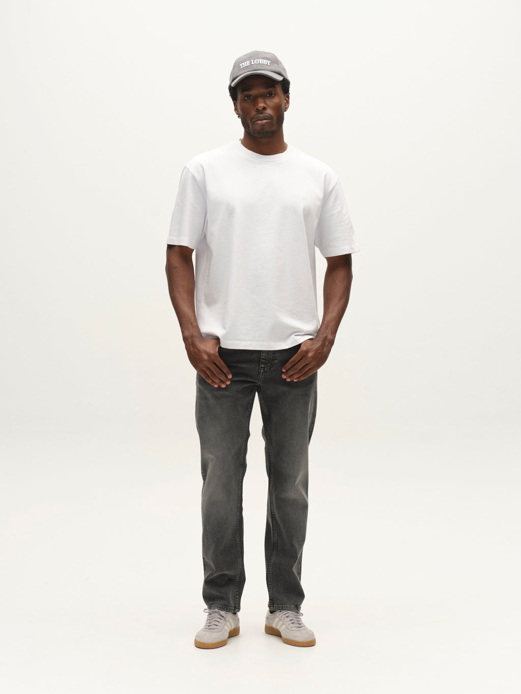 A man stands facing forward wearing a gray cap, plain white t-shirt, and Gabba GABmarc Blerb WW0032 Jeans in grey denim crafted from comfort stretch material, paired with gray sneakers against a plain white background.