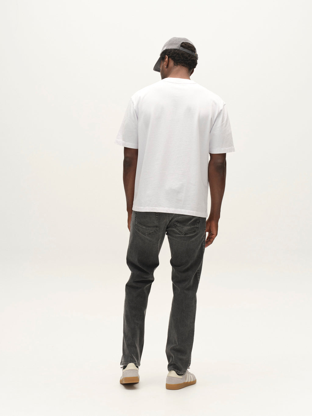 A man wearing a gray cap, white T-shirt, Gabba GABmarc Blerb WW0032 Jeans in grey denim crafted from comfort stretch fabric, and beige sneakers stands facing away in a minimal studio setting with a light background.