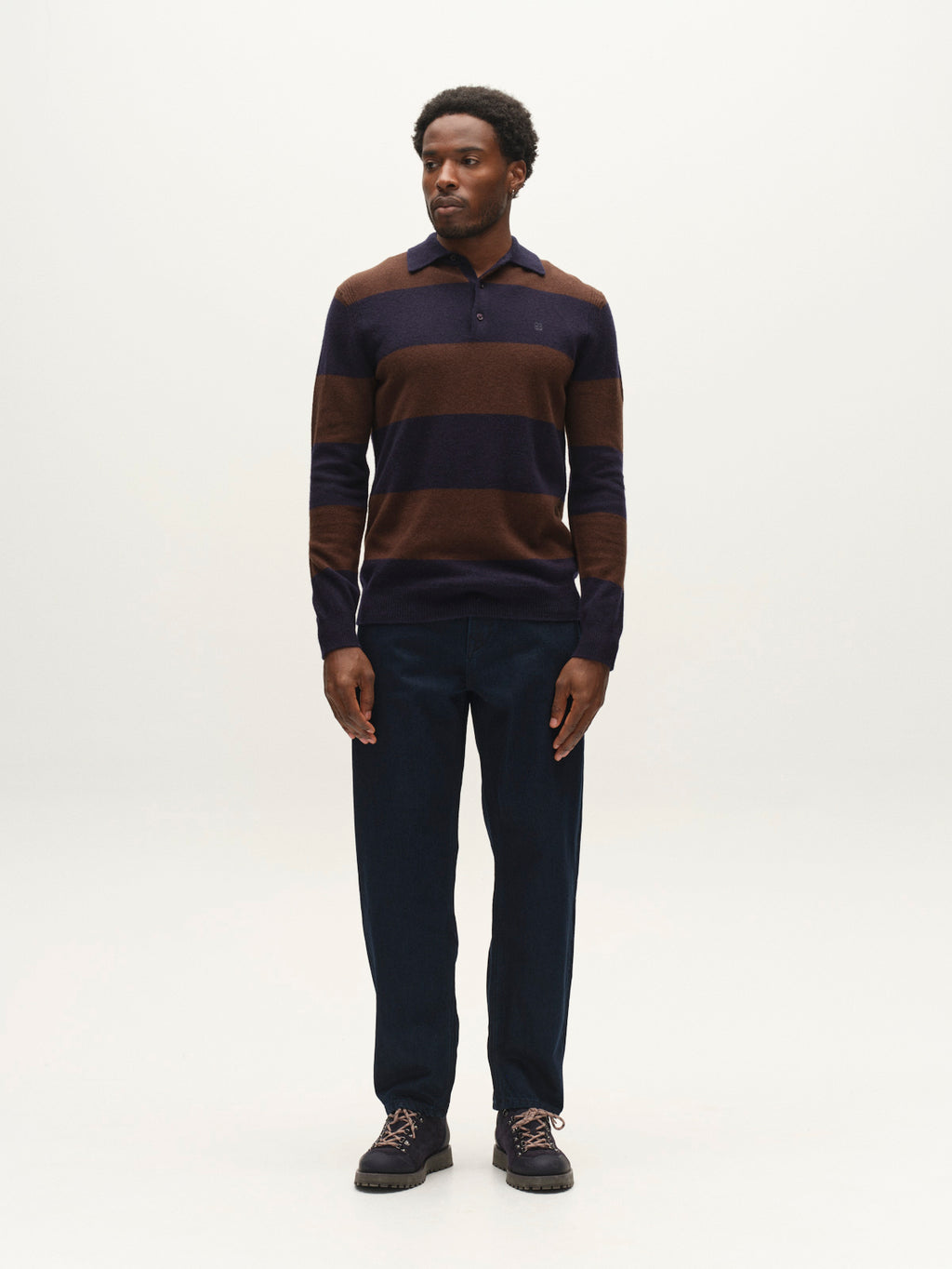 A man stands against a plain light background, wearing the Gabba GABworren Polo Stripes Aico LS Knit in Brown Stripe, styled as a long-sleeve polo. He pairs it with dark blue pants and dark shoes, facing forward with his hands at his sides.