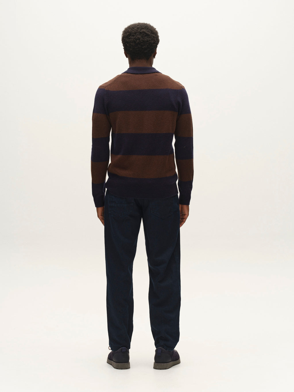 A person stands facing away, wearing the Gabba GABworren Polo Stripes Aico LS Knit in Brown Stripe, paired with dark pants and dark shoes, against a plain light background.