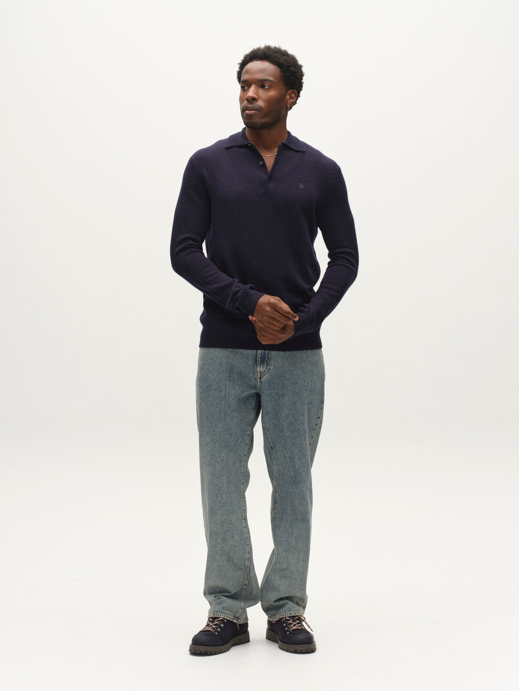 A man stands against a plain white background wearing the Gabba GABworren Polo Aico LS Knit in navy, paired with light blue jeans and black shoes, his hands clasped in front.