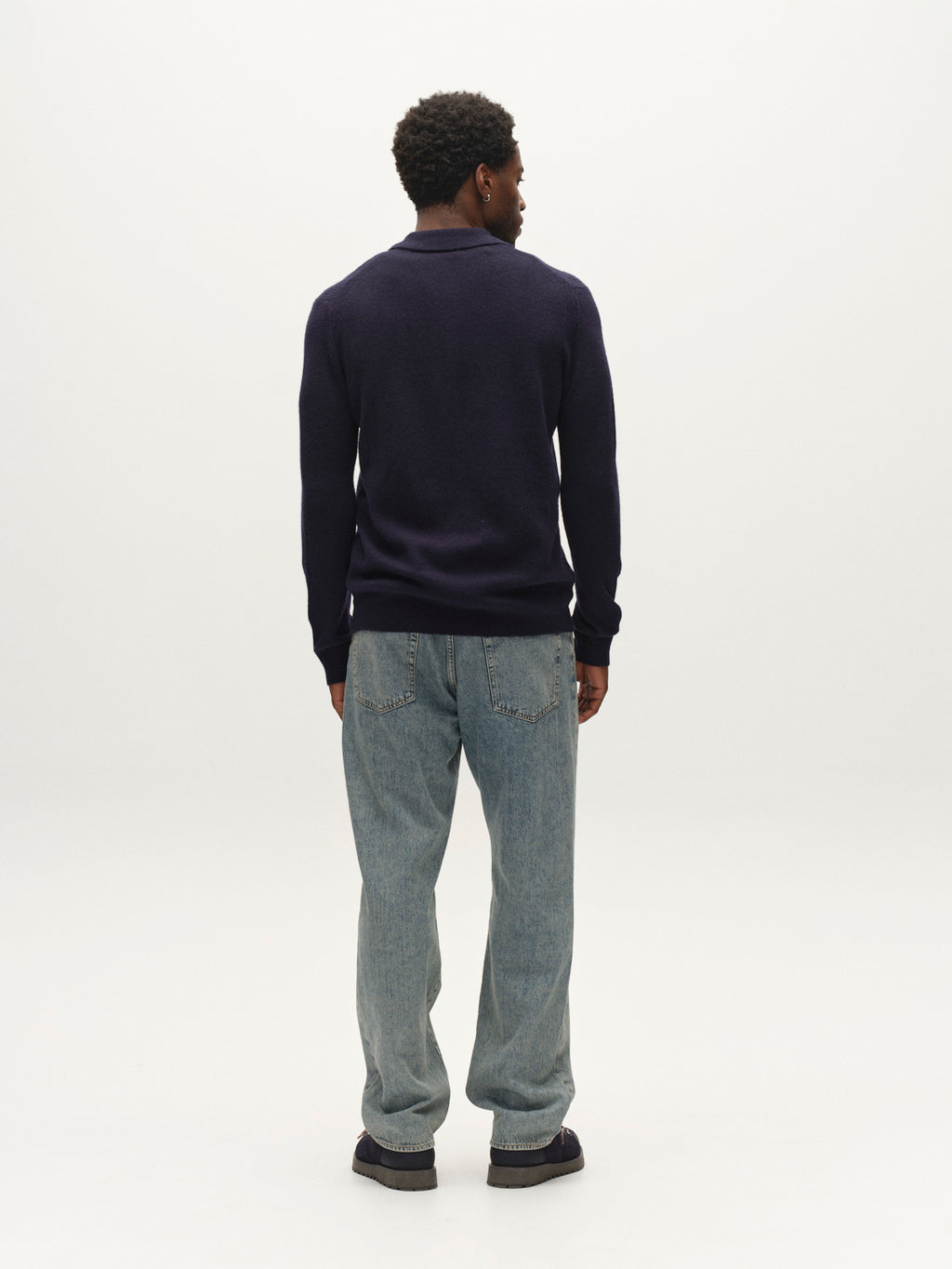 A person with short curly hair stands facing away in a plain white studio, wearing the Gabba GABworren Polo Aico LS Knit sweater in navy and light blue jeans.