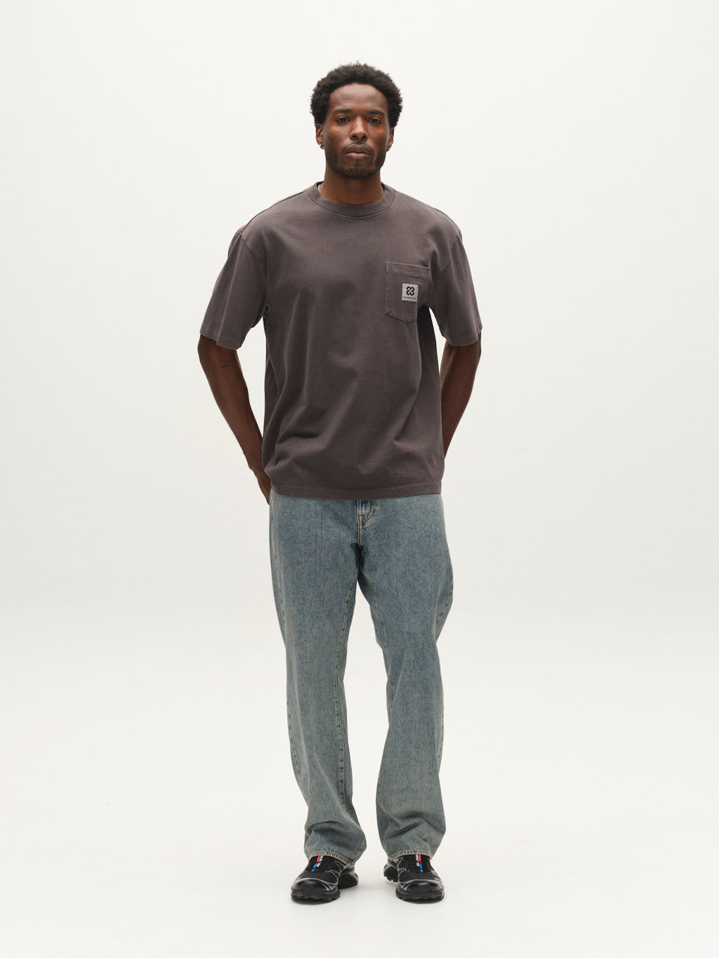 A man stands facing forward against a plain white background, wearing the Gabba GABnigel Wix Pkt Boxy Hour SS T-Shirt in dark grey—a loose, heavyweight cotton boxy tee with a chest pocket—paired with light blue jeans and black shoes.
