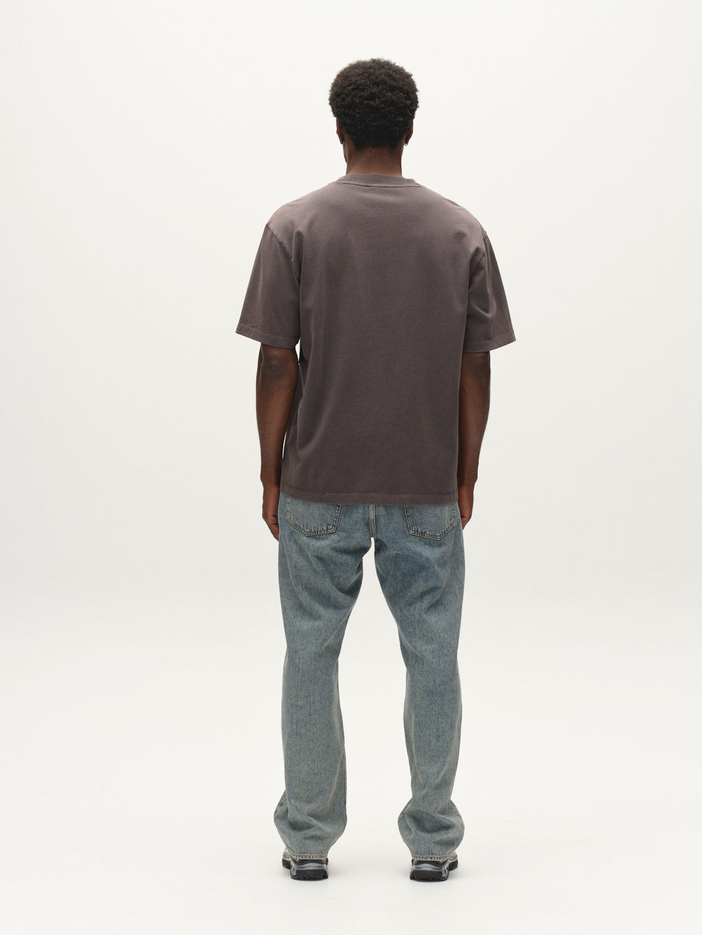 A person stands facing away, wearing the Gabba GABnigel Wix Pkt Boxy Hour SS T-Shirt in dark grey—a boxy heavyweight cotton tee—paired with light blue jeans and sneakers against a plain white background.