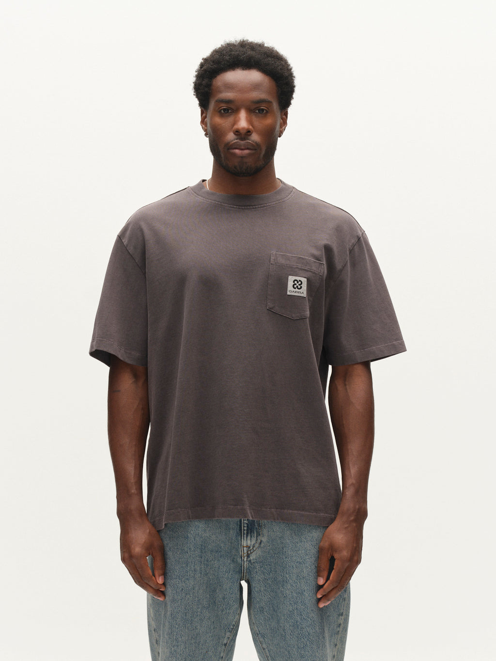 A man stands against a plain background, wearing the Gabba GABnigel Wix Pkt Boxy Hour SS T-Shirt in dark grey—a loose-fitting, boxy heavyweight cotton tee with a chest pocket—paired with light blue jeans. A subtle Gabba Brand Mark completes the look.