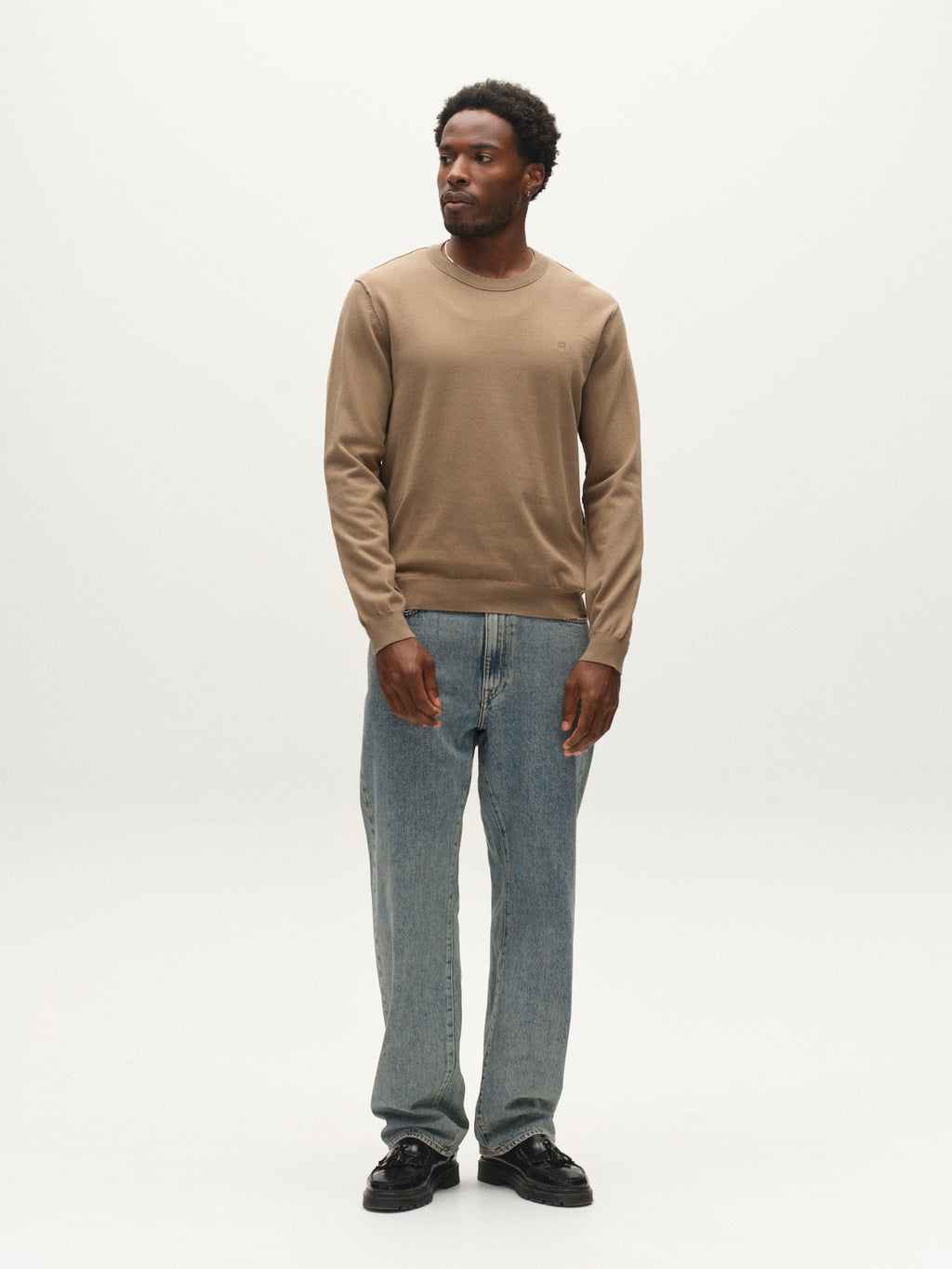 A man stands against a white background wearing the Gabba GABfumo Element Spy LS Knit sweater in Sand Crocker, paired with blue jeans and black shoes. He gazes slightly to the side with his arms relaxed at his sides.