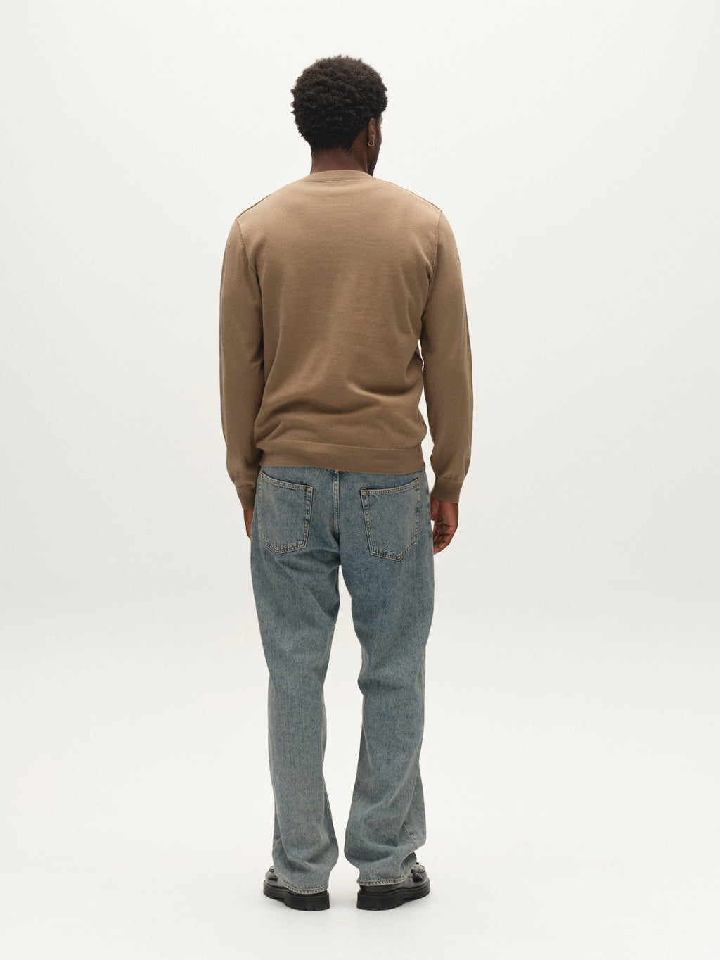 A man stands facing away from the camera, wearing the Gabba GABfumo Element Spy LS Knit - Sand Crocker, paired with blue jeans and black shoes against a plain white background.