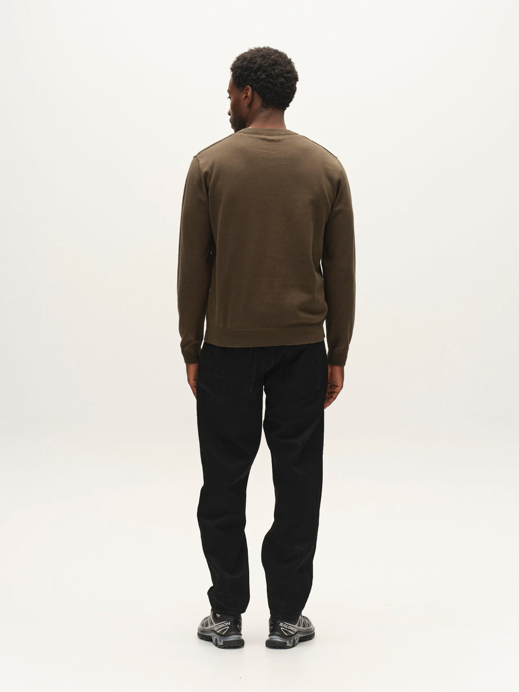 A man stands facing away from the camera, wearing the Gabba GABfumo Element Spy LS Knit in Forest Night—a regular fit long-sleeve shirt with a classic crew neck—paired with black pants and black sneakers, against a plain white background.