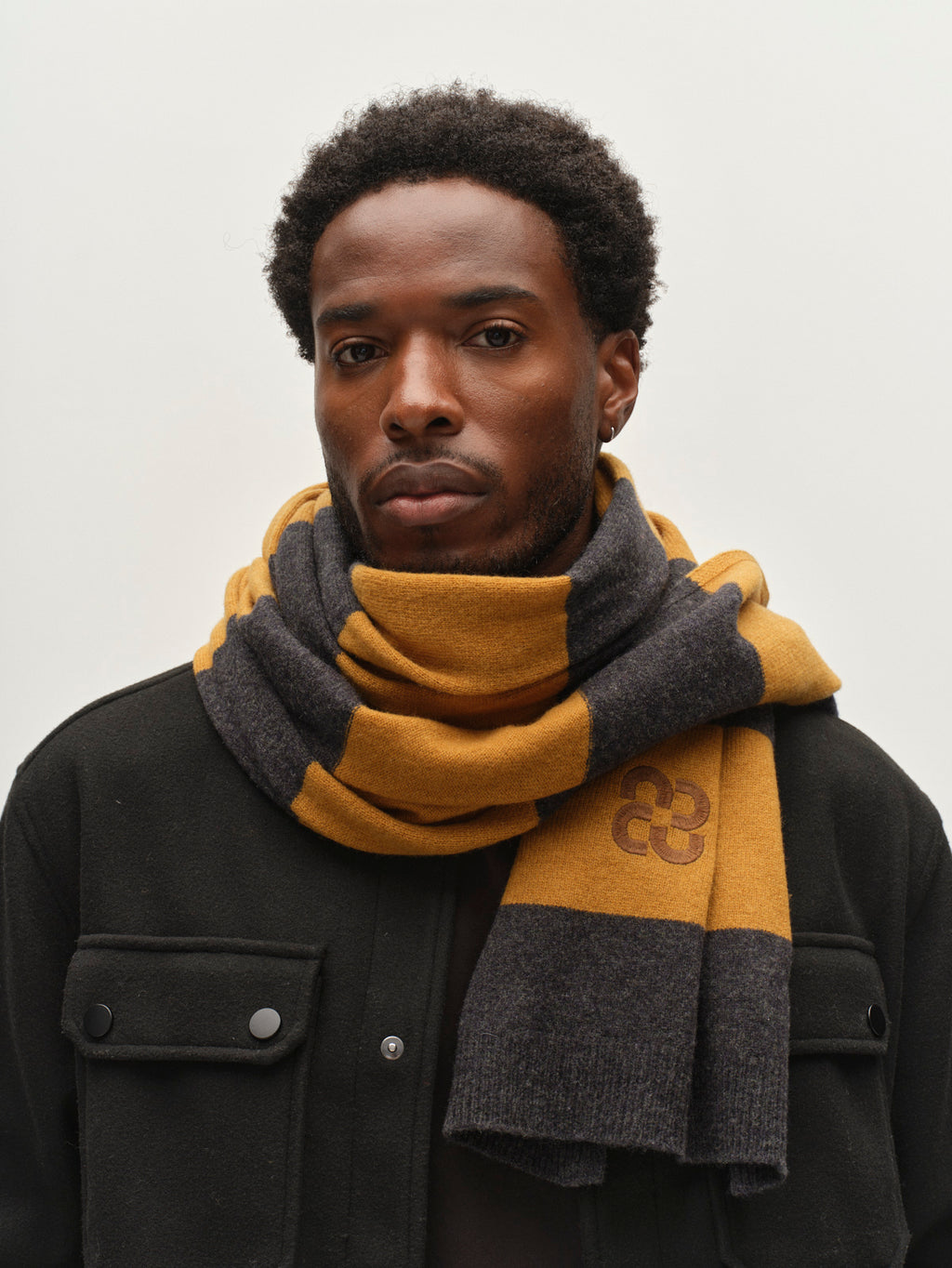 A man wearing a black jacket and the Gabba GABwest Aico Scarf – Yellow Stripes, made from merino wool with bold yellow and gray stripes, stands against a plain light background, looking directly at the camera with a neutral expression. This cozy Gabba accessory adds both warmth and style to his look.