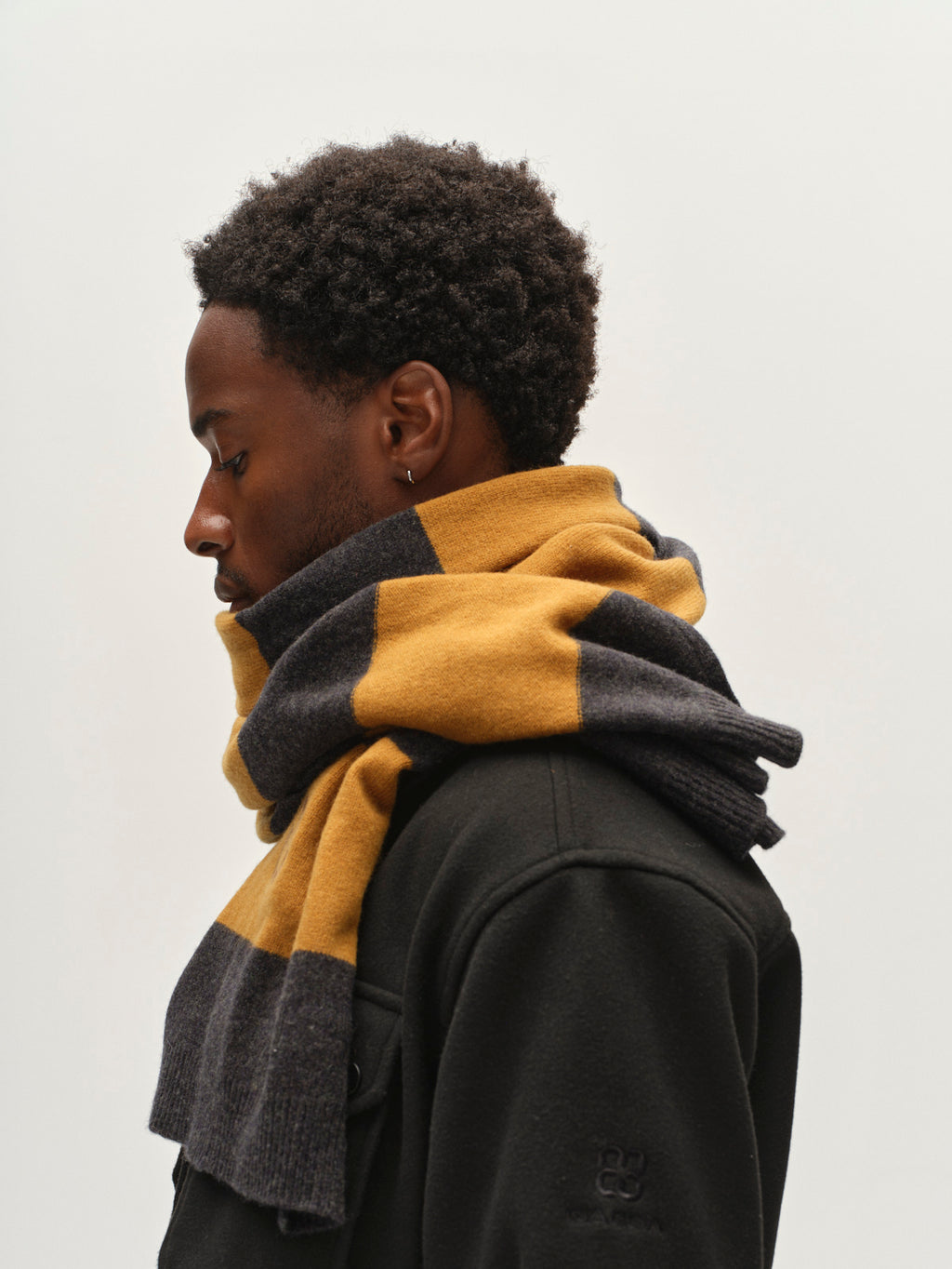 A person with short curly hair, shown in profile against a plain light background, wears a black coat and the Gabba GABwest Aico Scarf in Yellow Stripes—a stylish merino wool winter accessory.