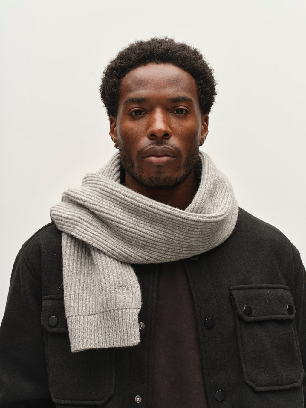 A man with short curly hair wearing a black jacket and the Gabba GABeast Flash Scarf in Grey Mel—a thick, rib-knit wool scarf—stands against a plain white background, showcasing this stylish winter accessory.