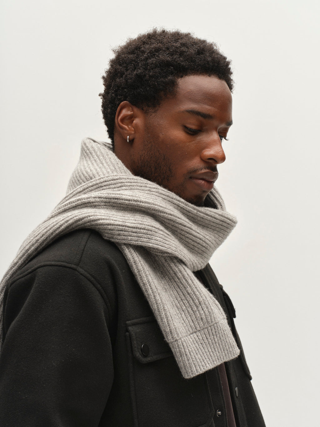 A man with short curly hair wears the Gabba GABeast Flash Scarf in Grey Mel, paired with a black jacket, looking down against a plain light background.