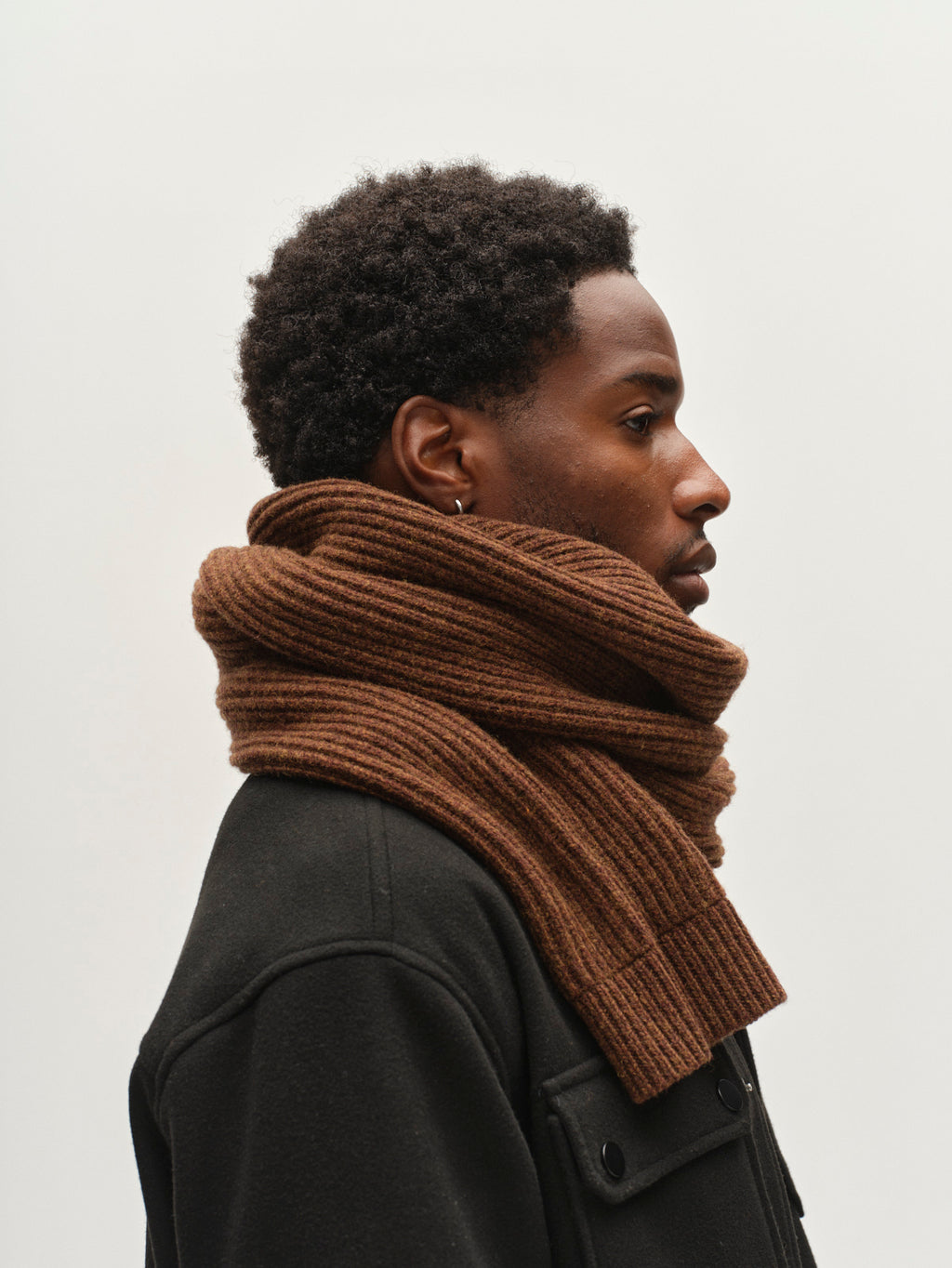A person with short curly hair wears the Gabba GABeast Flash Scarf in Brown Slate—a thick, rib-knit wool scarf—wrapped around their neck with a dark coat, facing right against a plain white background; it’s the perfect winter accessory.